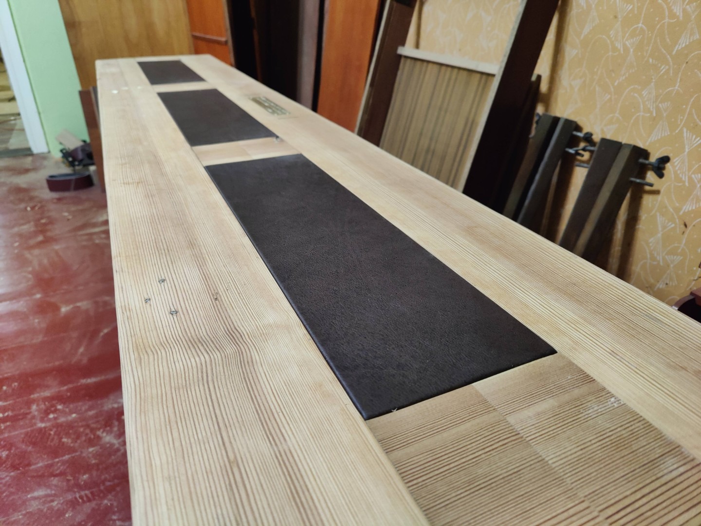 Długi, jasny blat drewniany z ciemnymi wstawkami, widoczne ślady obróbki, surowe wykończenie, w tle warsztat stolarski z elementami drewna i narzędziami.