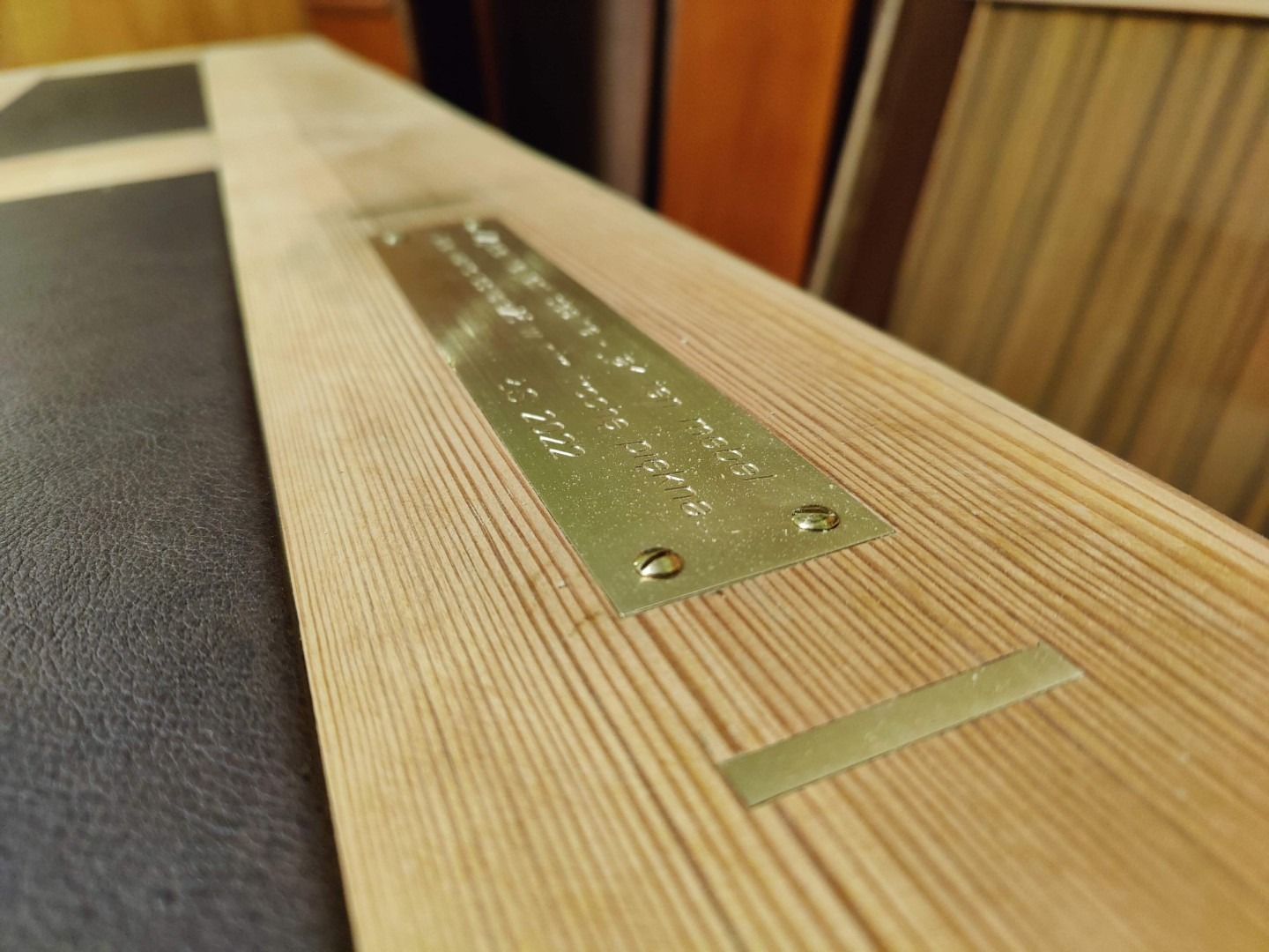 Detal drewnianego blatu z metalową tabliczką z grawerem, widoczne słoje drewna i fragment skórzanego wykończenia. Ujęcie z bliska.