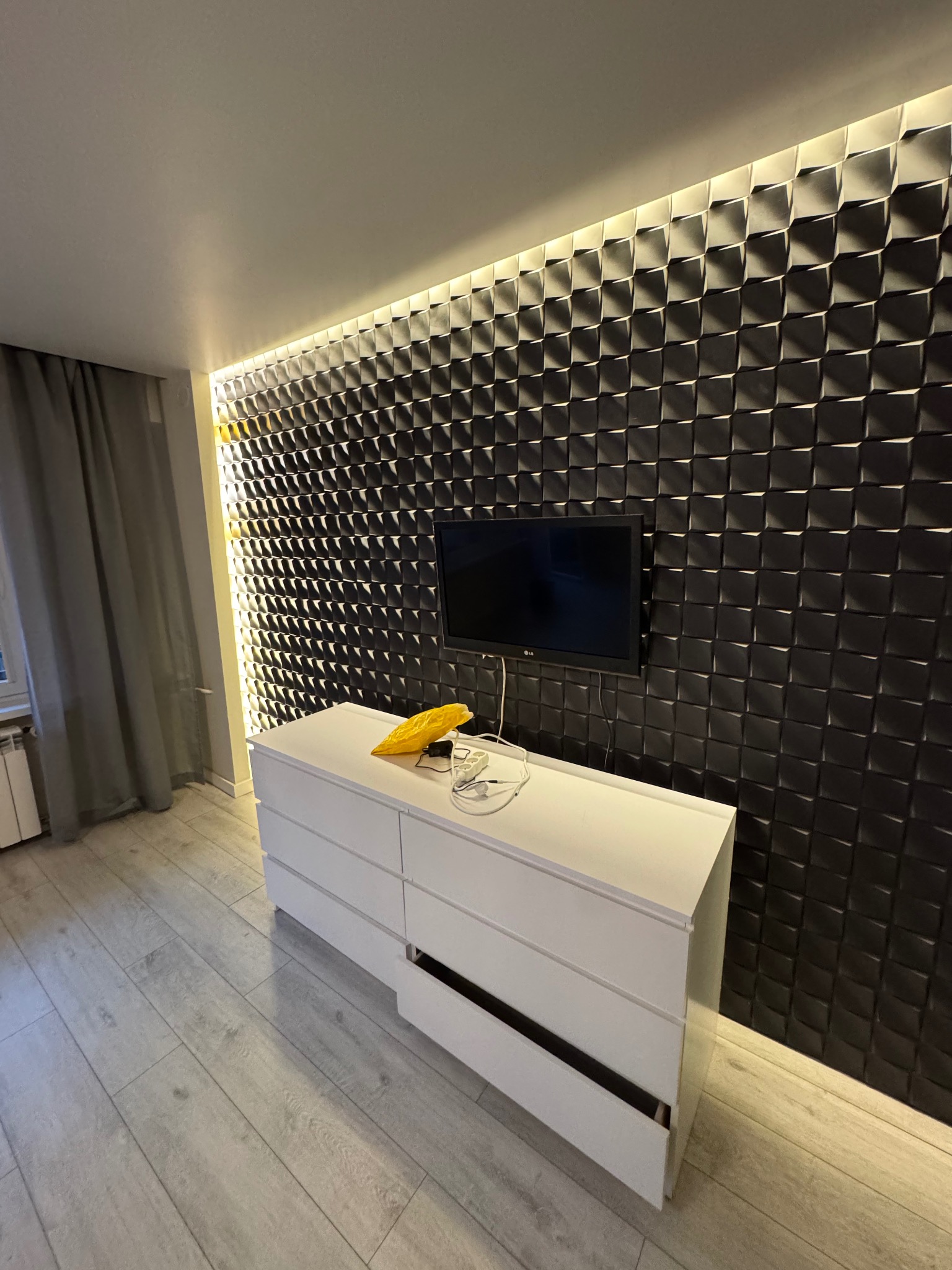 Nowoczesne wnętrze w Olkuszu z dekoracyjną ścianą 3D, podświetlaną listwą LED i białymi panelami podłogowymi, uzupełnione białą komodą z otwartą szufladą i telewizorem.
