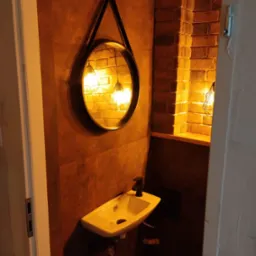 Umywalka z czarną baterią i okrągłe lustro w czarnej ramie na tle ściany z płytek w odcieniach brązu i ceglanej wnęki z oświetleniem w małej łazience.