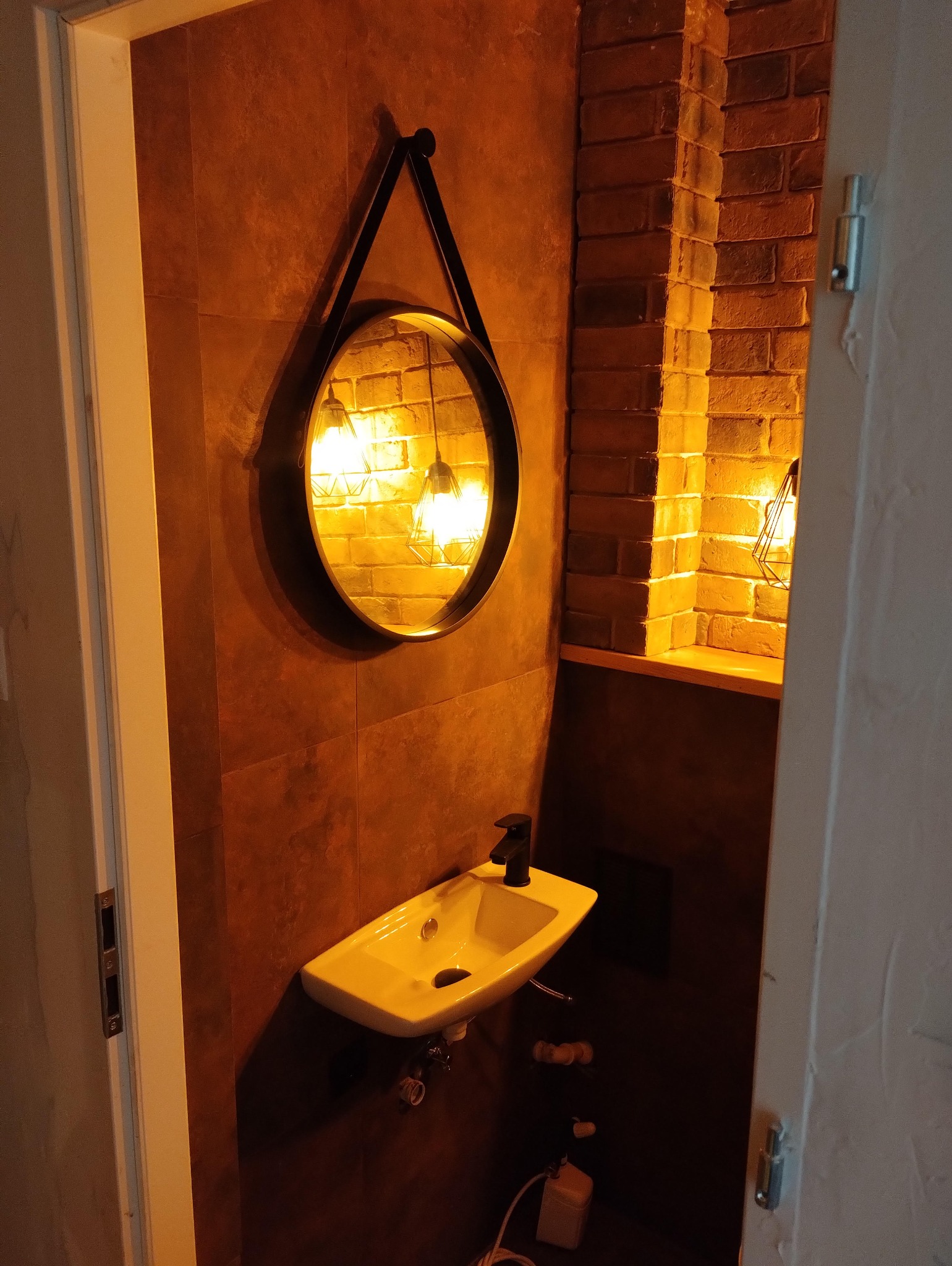 Umywalka z czarną baterią i okrągłe lustro w czarnej ramie na tle ściany z płytek w odcieniach brązu i ceglanej wnęki z oświetleniem w małej łazience.