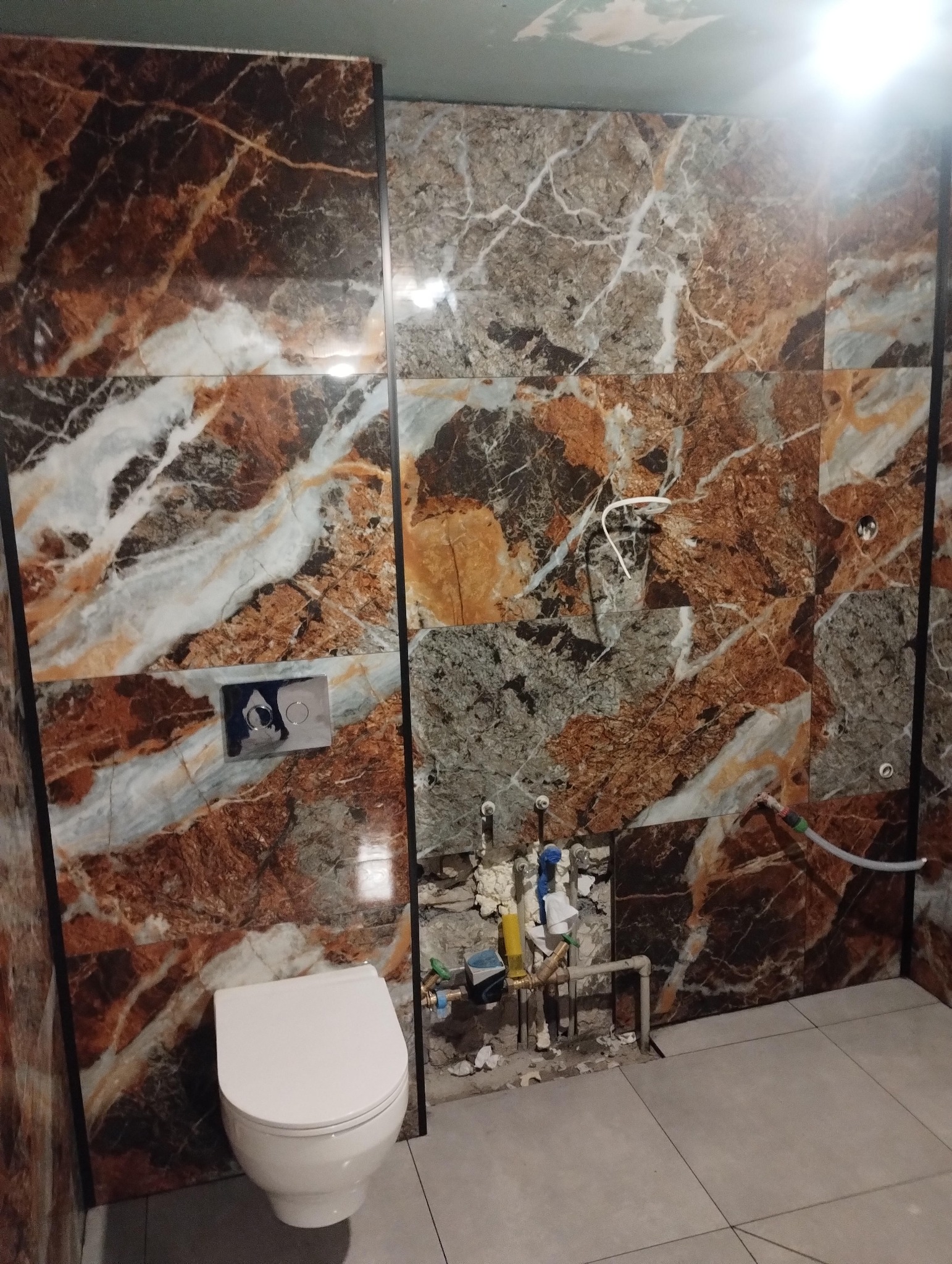 Łazienka w trakcie wykańczania: marmurowe płytki ścienne w odcieniach brązu i szarości, biała toaleta, widoczne instalacje wodne przed zabudową.