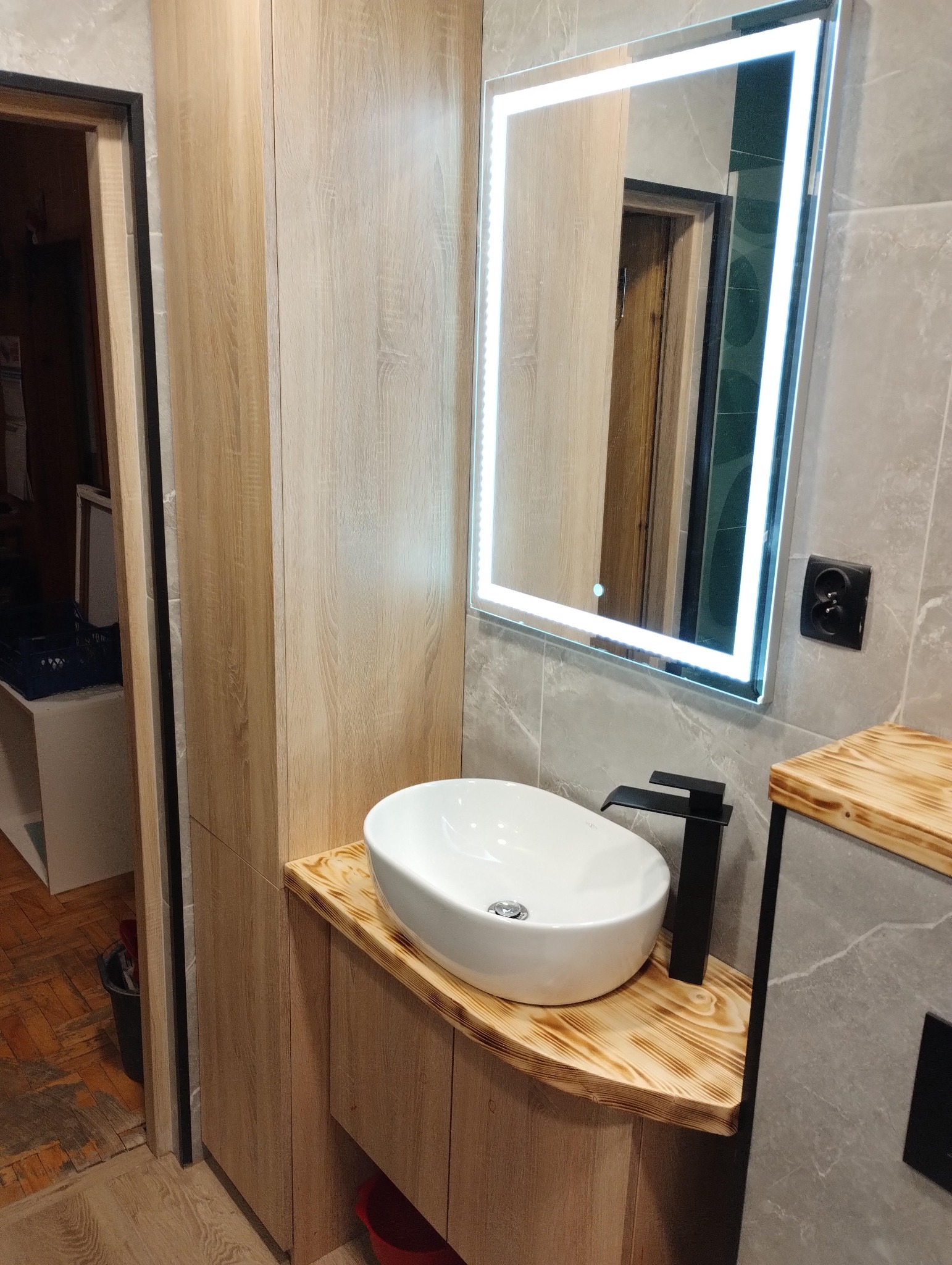 Nowoczesna łazienka z umywalką nablatową, szafką z drewnopodobnym fornirem i lustrem z podświetleniem LED. Szare płytki imitujące marmur na ścianie.