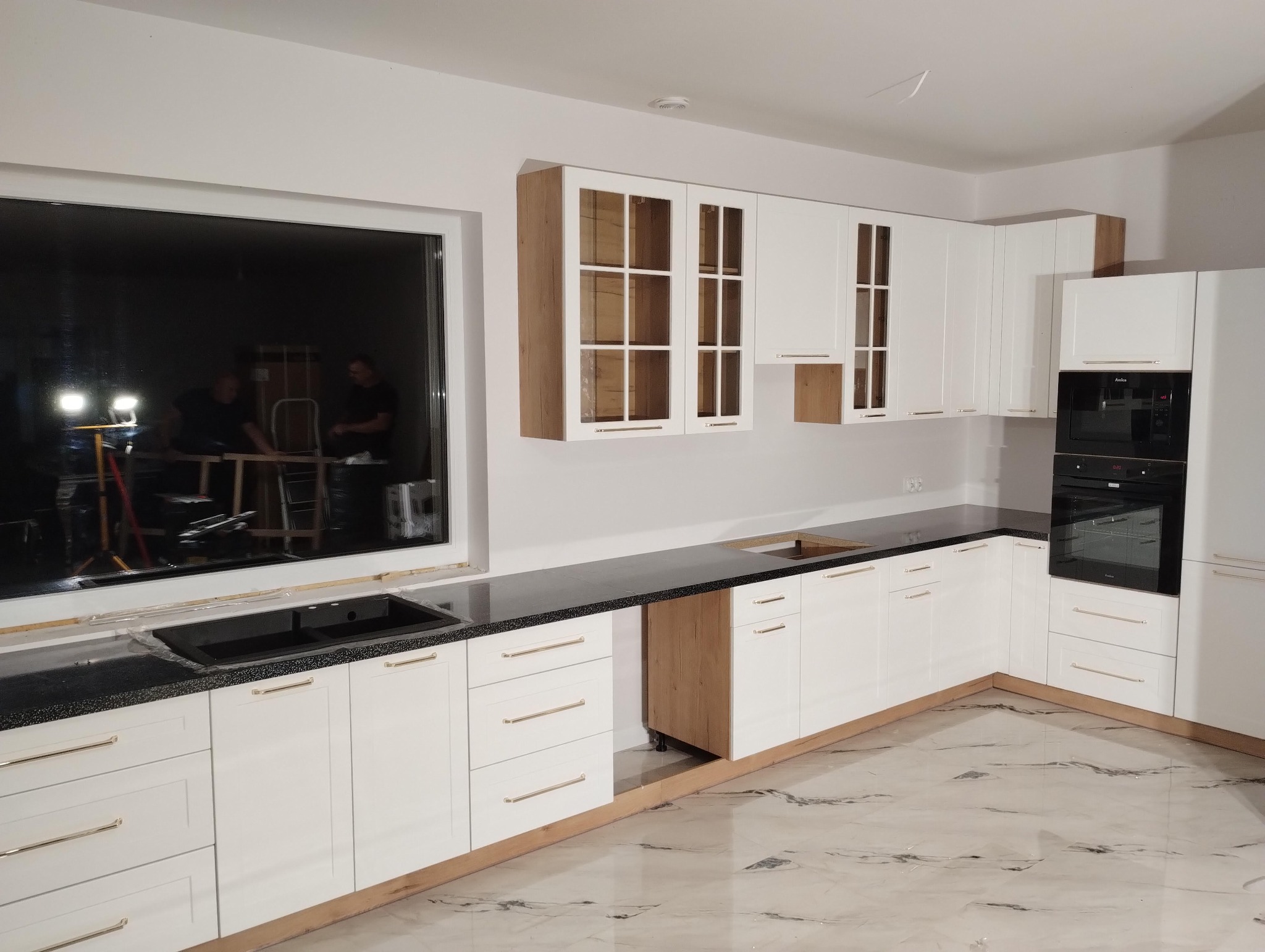 Nowoczesna kuchnia z białymi szafkami, czarnym blatem i marmurową podłogą. W oknie widoczne narzędzia i ekipa montażowa. Górne szafki z elementami drewna.