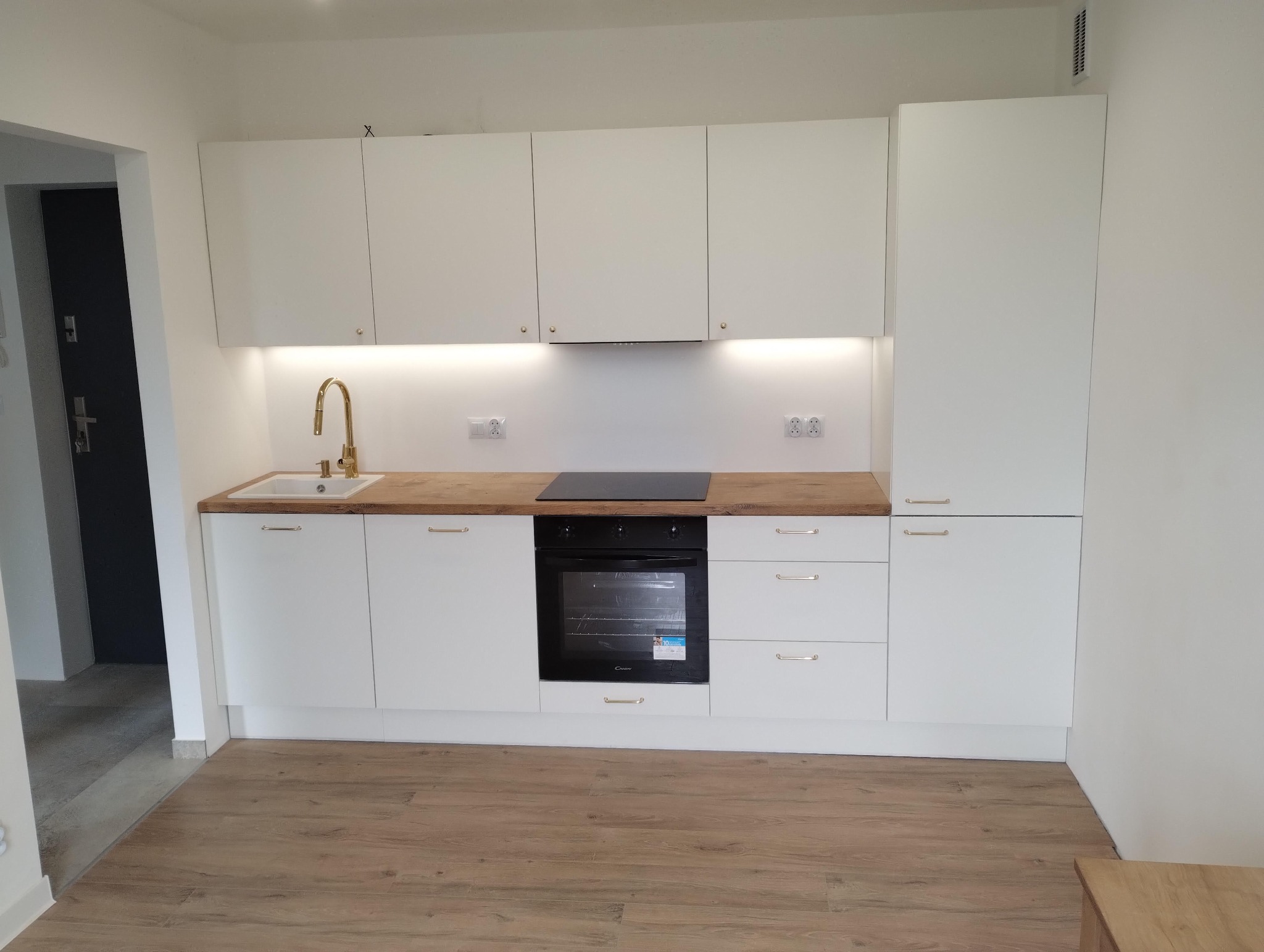 Biała, minimalistyczna zabudowa kuchenna z drewnianym blatem i złotą armaturą. Widok na piekarnik i płytę indukcyjną. Podświetlenie szafek.