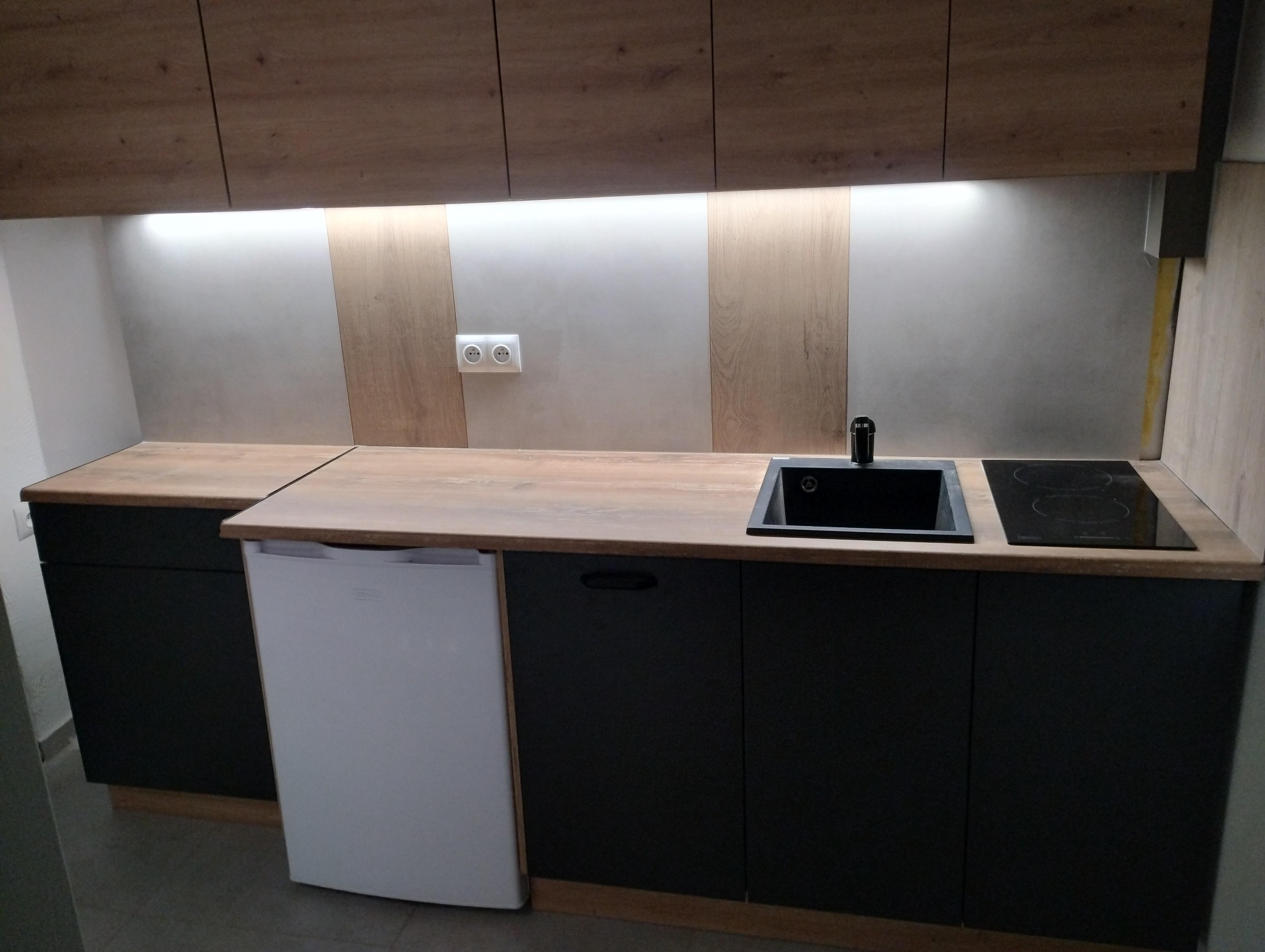 Nowoczesna kuchnia: czarne szafki, drewniany blat, biała lodówka. Minimalistyczny design, oświetlenie LED pod szafkami, płyta indukcyjna i czarny zlew.