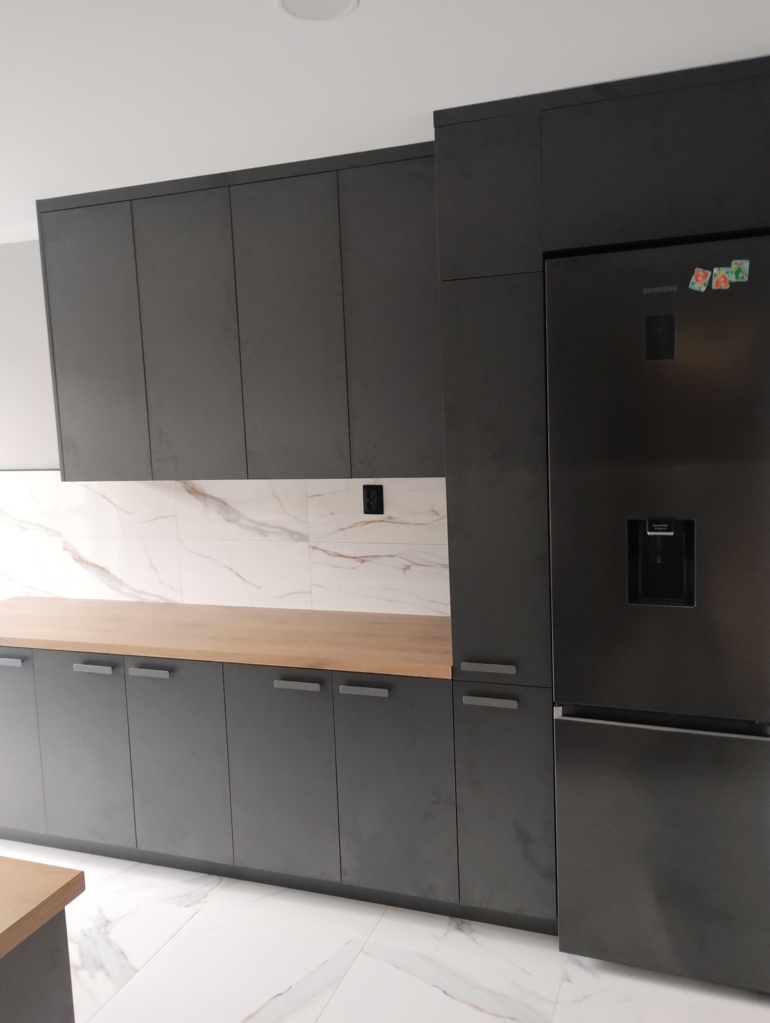 Nowoczesna kuchnia w odcieniach szarości z drewnianym blatem i marmurowymi płytkami na ścianie. Minimalistyczne fronty szafek, duża lodówka.