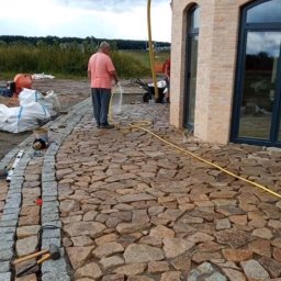 Art-Stone bruk - Ułożona z nieregularnych, brązowych kamieni ścieżka, obrzeżona szarą kostką granitową. Mężczyzna podlewa powierzchnię. Widoczne narzędzia brukarskie.