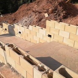 Art-Stone bruk - Budowa muru oporowego z pustaków betonowych, zbrojenie widoczne w pustakach. W tle ziemia.