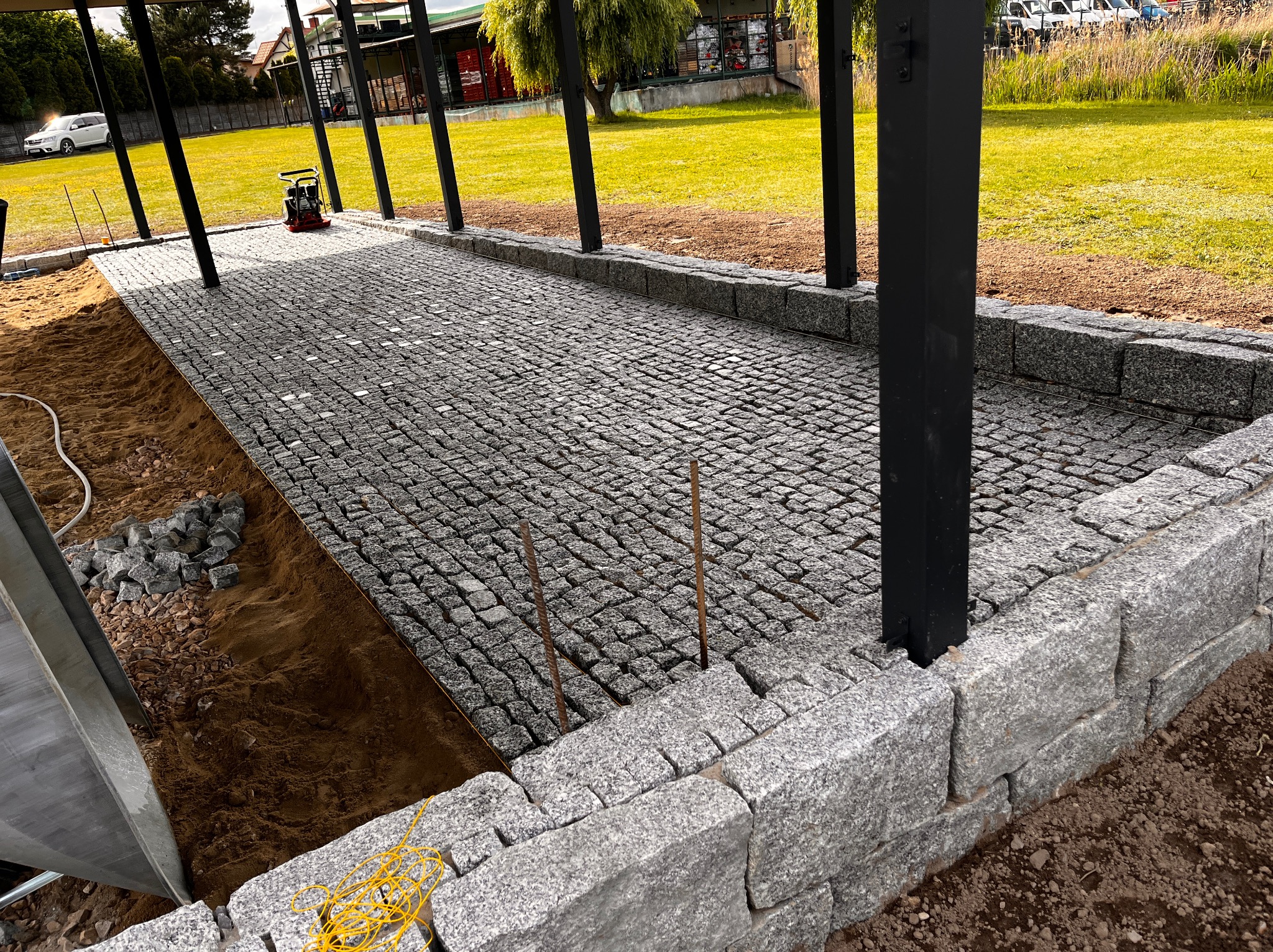 Układanie kostki granitowej w Głownie. Widoczna część ułożonej nawierzchni z kostki, obrzeża i przygotowany teren pod dalsze prace. W tle sprzęt budowlany.