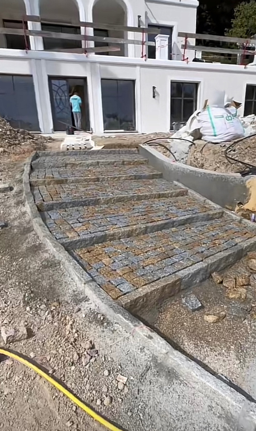 Schody z kostki granitowej prowadzące do białego domu. Widoczny etap prac, z żółtym wężem ogrodowym w tle. Kostka w odcieniach szarości i beżu.