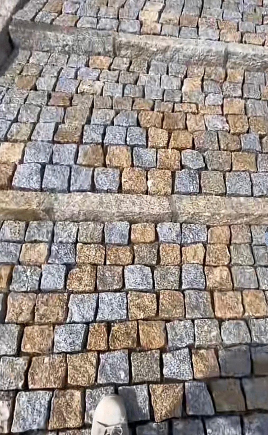 Ułożona kostka granitowa w odcieniach szarości i brązu tworząca schody. Widoczny fragment buta osoby stojącej na schodach. Perspektywa z góry.