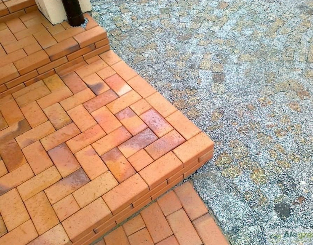 Schody i ścieżka z czerwonej kostki, ułożone w wzór jodełki, sąsiadujące z szarą, drobną nawierzchnią. Widok z góry, fragment rynny.