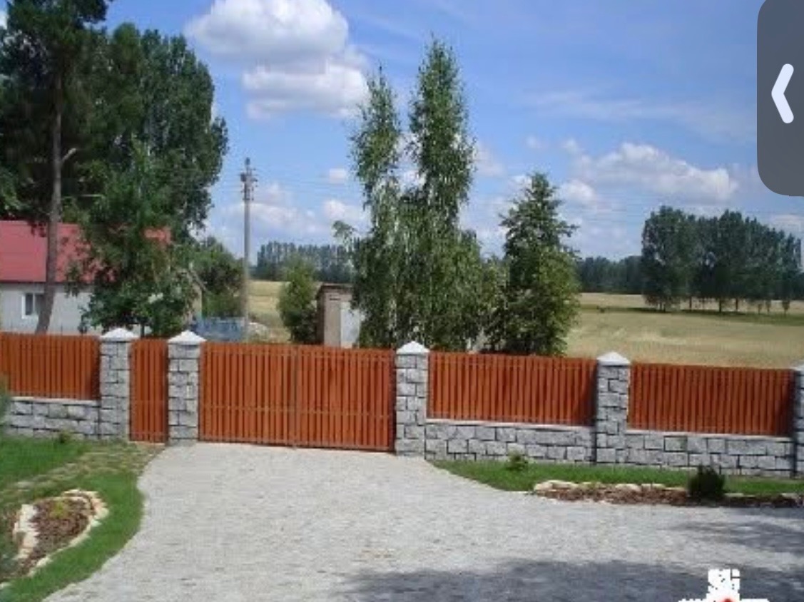 Drewniana brama wjazdowa z kamiennymi słupkami i podmurówką, prowadząca do domu. Widoczny podjazd z kostki i elementy zieleni. Słoneczny dzień.