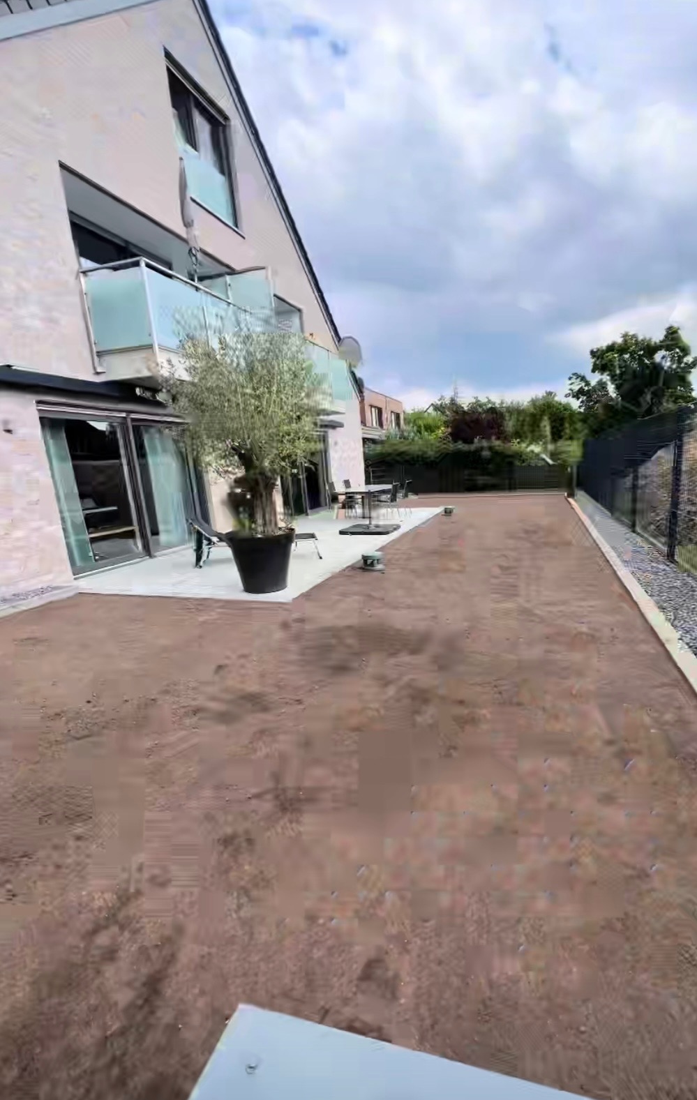 Przygotowany teren pod ogród przy nowoczesnym domu z tarasem i dużym drzewem w donicy. Widoczny fragment ogrodzenia i niebo z chmurami.