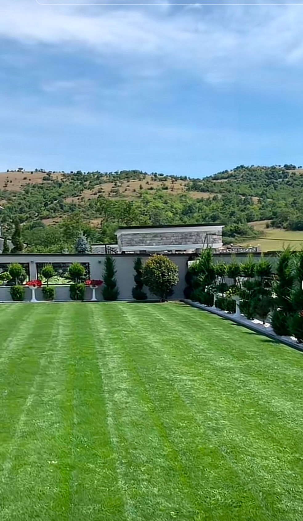 Perfekcyjnie utrzymany trawnik z równo przystrzyżoną trawą i ozdobnymi nasadzeniami na tle wzgórz i błękitnego nieba. Widoczne pasy po kosiarce.