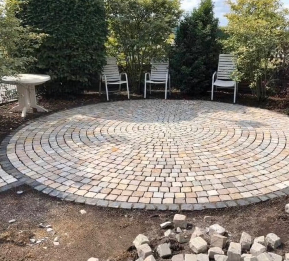 Okrągły plac z kostki granitowej w ogrodzie, otoczony zielenią i trzema białymi krzesłami. Widoczne niedokończone fragmenty i kostka obok.