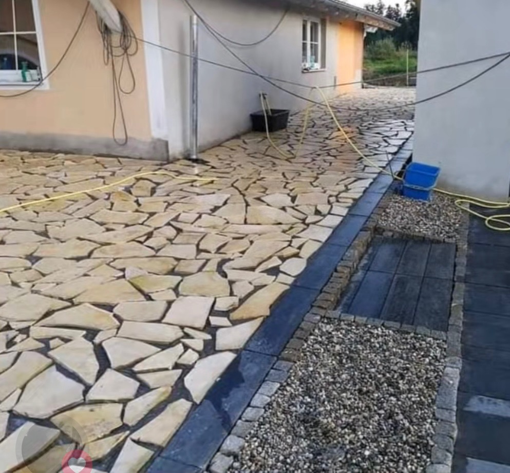 Ułożona ścieżka z nieregularnych, płaskich kamieni w odcieniach beżu. Obok widoczny drenaż z czarnych elementów i kamieni ozdobnych. W tle budynek.