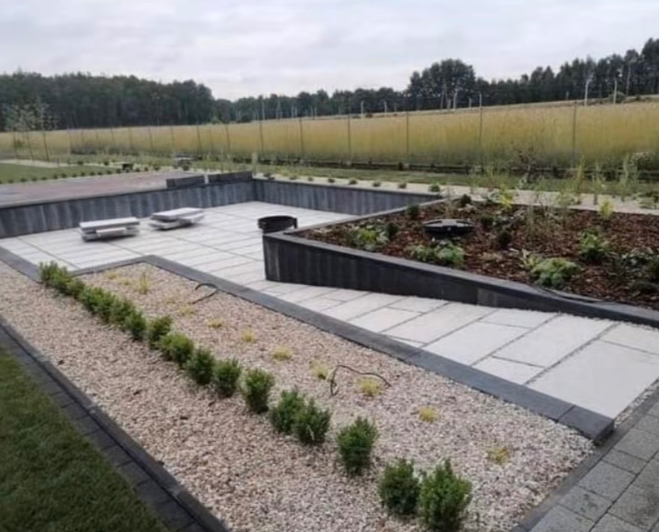Nowoczesny ogród z geometrycznym układem kostki, rabatami z korą i drobnym żwirem. Widoczne małe krzewy i trawy ozdobne na tle łąki i drzew.