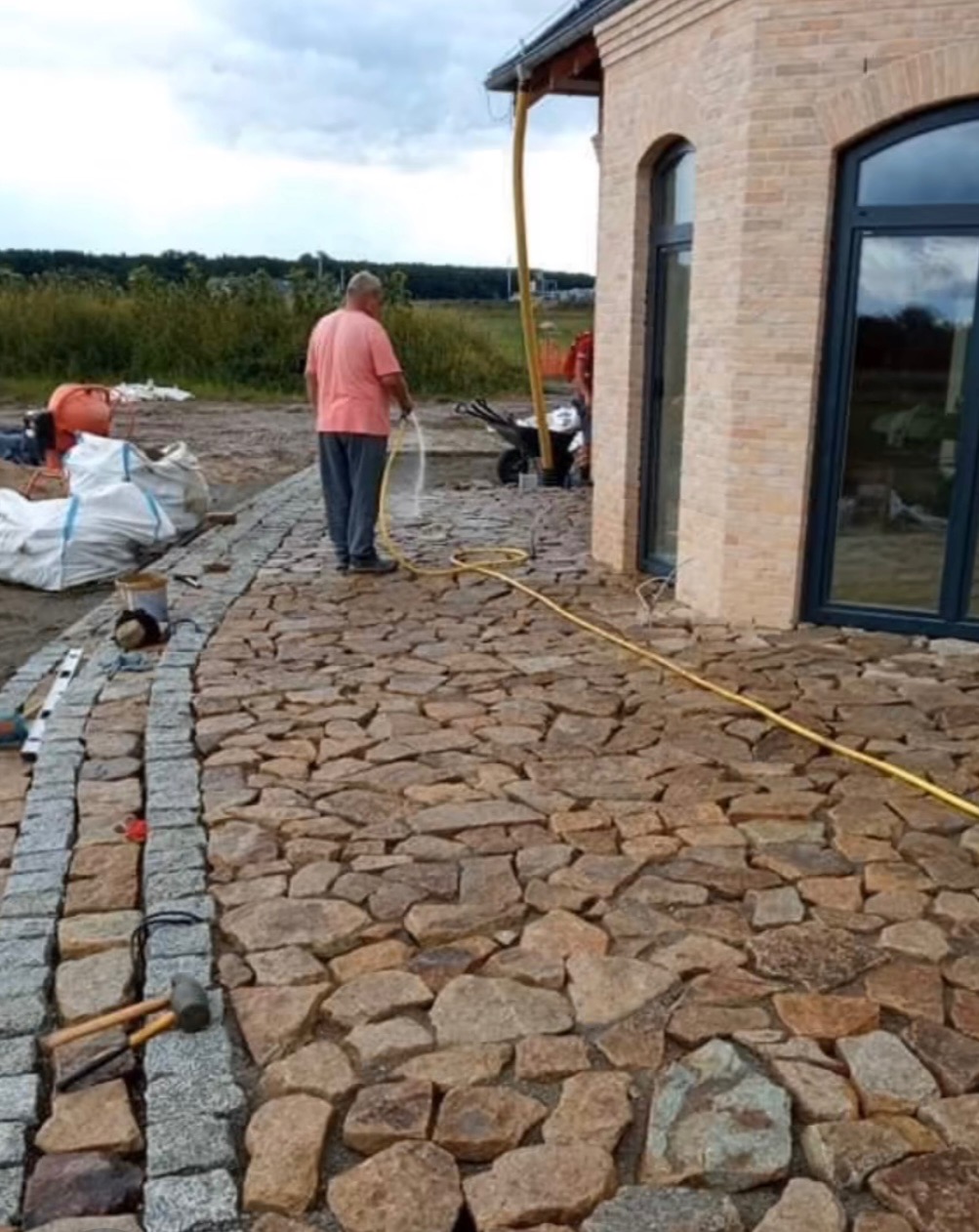 Ułożona z nieregularnych, brązowych kamieni ścieżka, obrzeżona szarą kostką granitową. Mężczyzna podlewa powierzchnię. Widoczne narzędzia brukarskie.