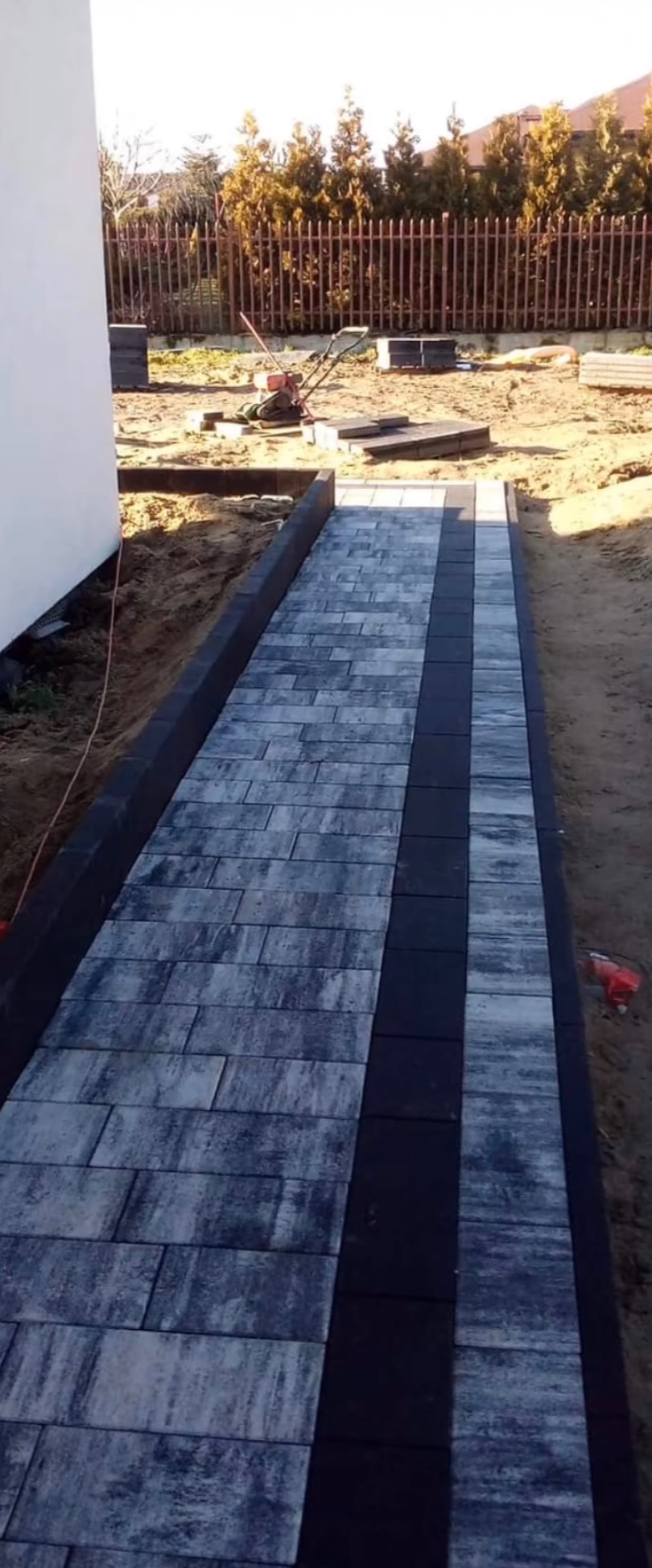 Ułożona ścieżka z kostki brukowej w odcieniach szarości, z ciemnym pasem pośrodku, obrzeża z kostki. W tle widoczny trawnik, ogrodzenie i krzewy.