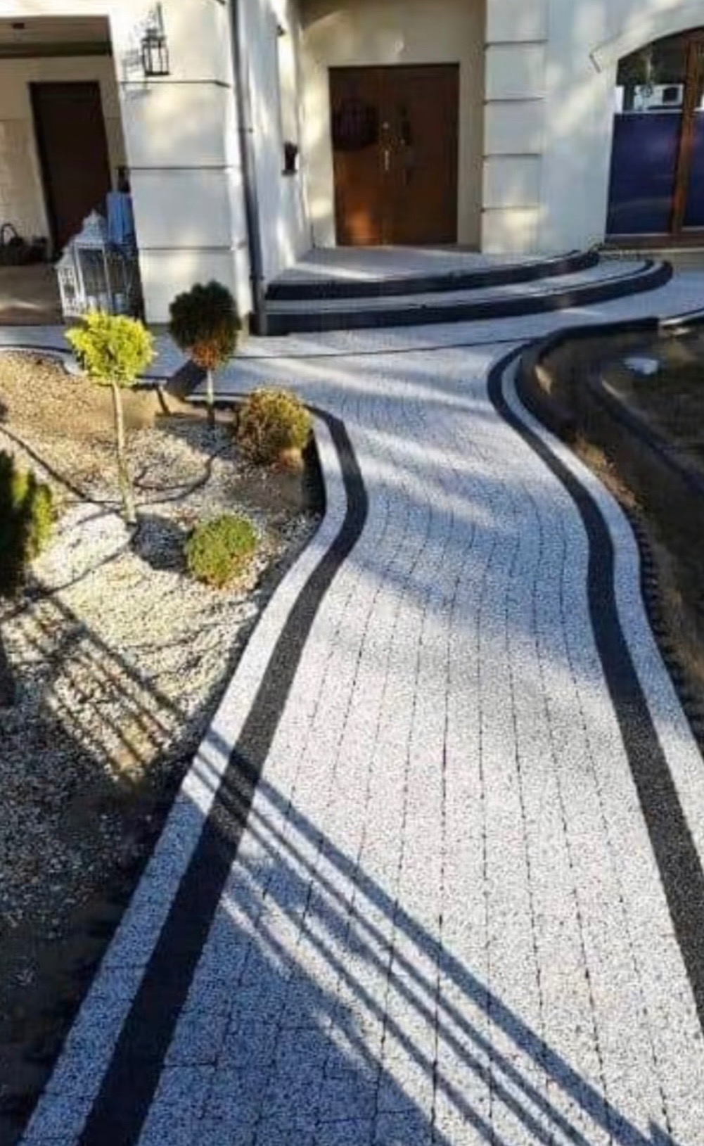 Kręty chodnik z jasnej kostki brukowej z czarnym obramowaniem prowadzi do domu. Widoczne małe drzewka i krzewy. Cień rzucany przez ogrodzenie.