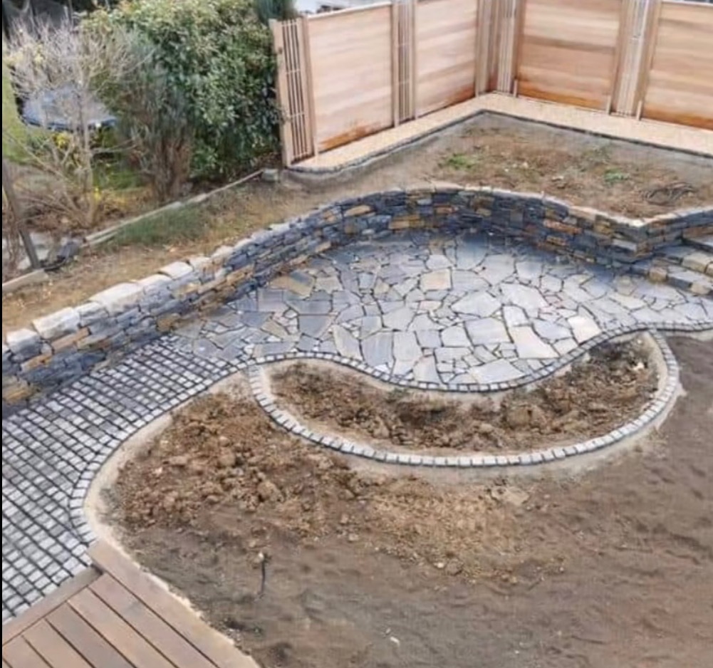 Aranżacja ogrodu z kamienną ścieżką i tarasem z nieregularnych płyt, otoczonych murkiem. W tle drewniane ogrodzenie, ziemia przygotowana pod nasadzenia.