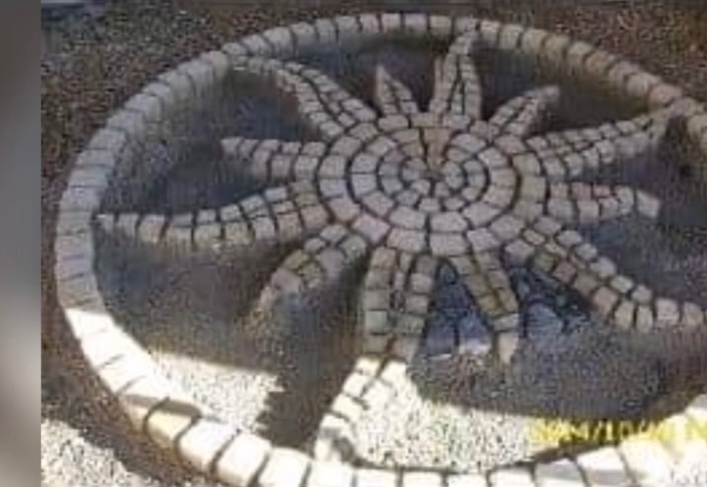 Okrągła kompozycja z kostki brukowej w kształcie słońca, otoczona obramowaniem z kostki. Dekoracyjny element w ogrodzie, wykonany z jasnoszarej kostki.