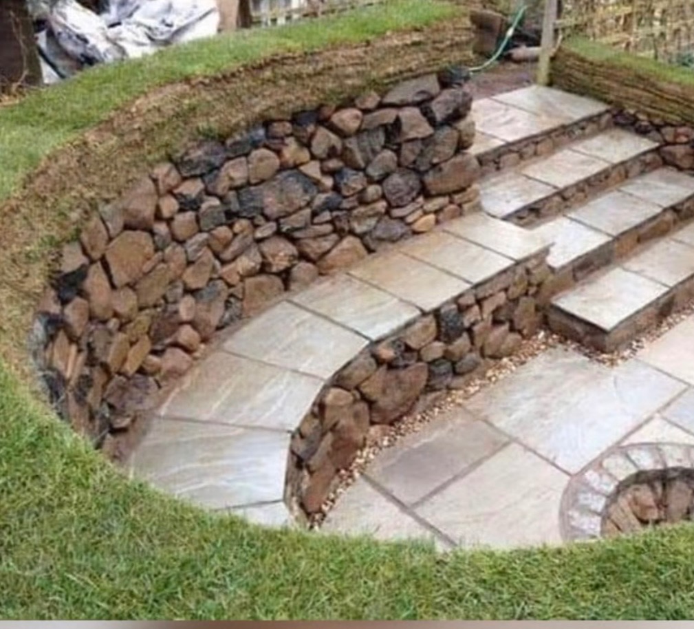 Kamienne schody ogrodowe z trawiastym obrzeżem i murem oporowym z kamienia polnego. Jasne płyty chodnikowe tworzą stopnie i ścieżkę.