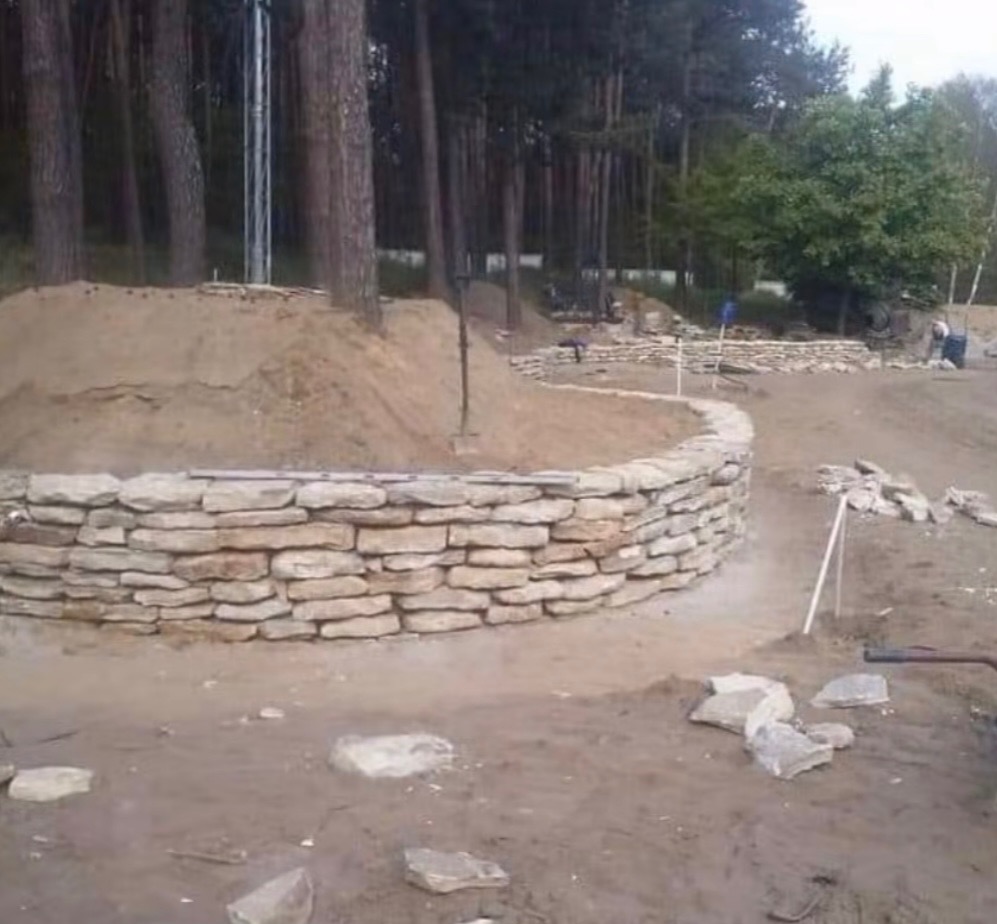 Budowa kamiennego murku oporowego z piaskowca na terenie zalesionym. W tle widać kopiec piasku i kontynuację prac. Częściowo ukończona konstrukcja.