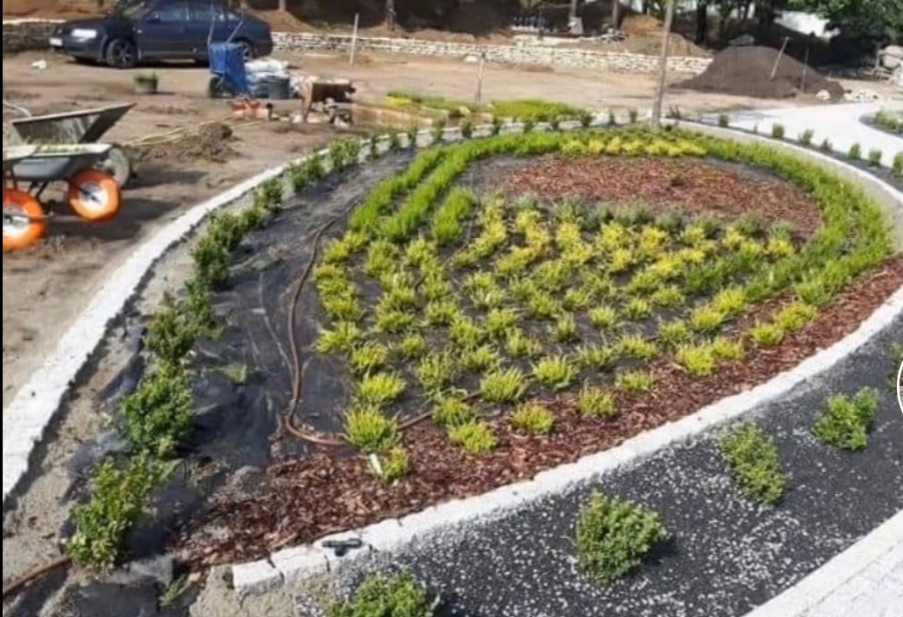 Aranżacja ogrodu z roślinami w geometrycznym wzorze, obrzeża z kostki, w tle wózek i auto. Dekoracyjny ogród z elementami nasadzeń i utwardzonej nawierzchni.