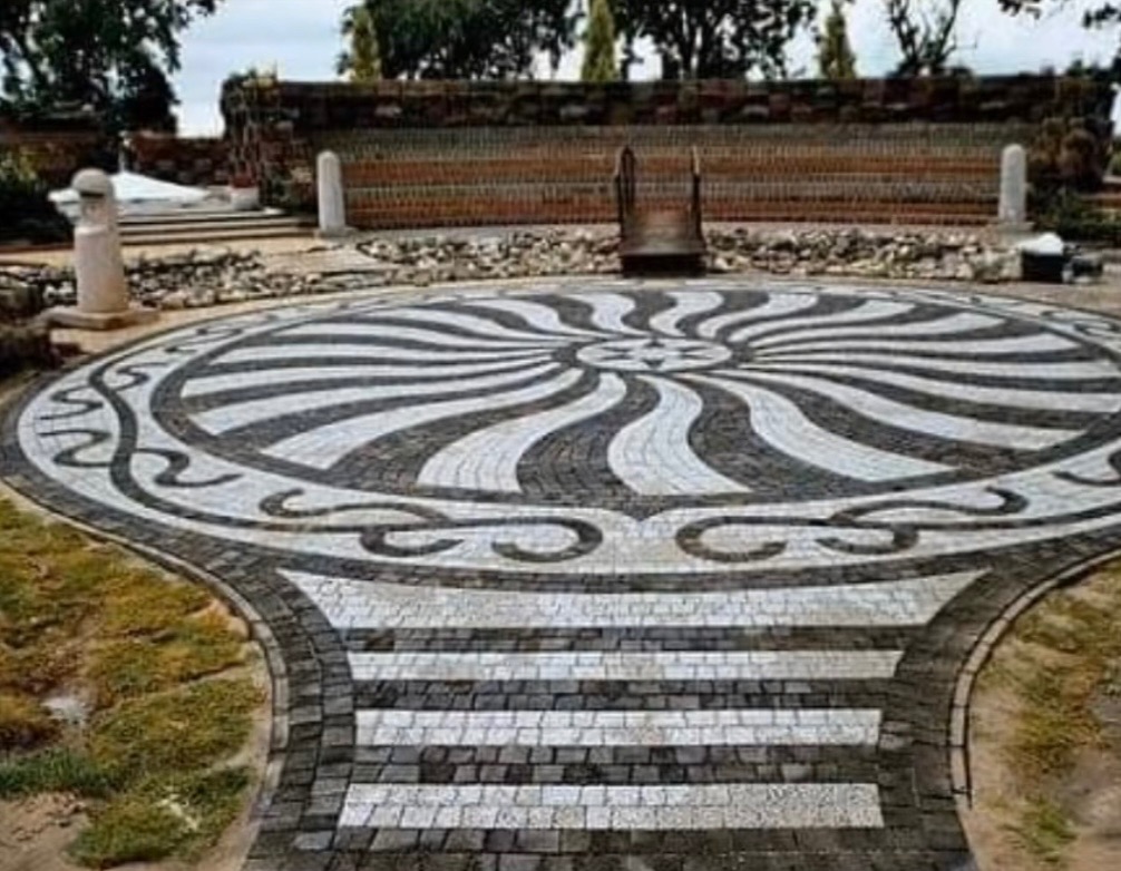 Okrągła, dekoracyjna nawierzchnia z kostki granitowej w odcieniach szarości i bieli, z motywem rozchodzących się promieni i ornamentami. Tło stanowi mur z cegły.