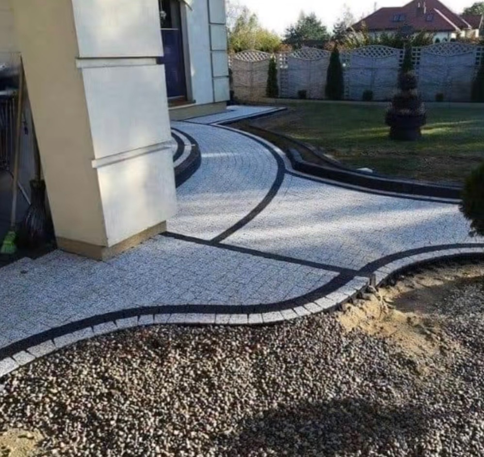 Kręta ścieżka z jasnej kostki granitowej z czarnymi obrzeżami prowadzi przez ogród. Widoczne luźne kamienie i trawnik. Ujęcie z poziomu gruntu.