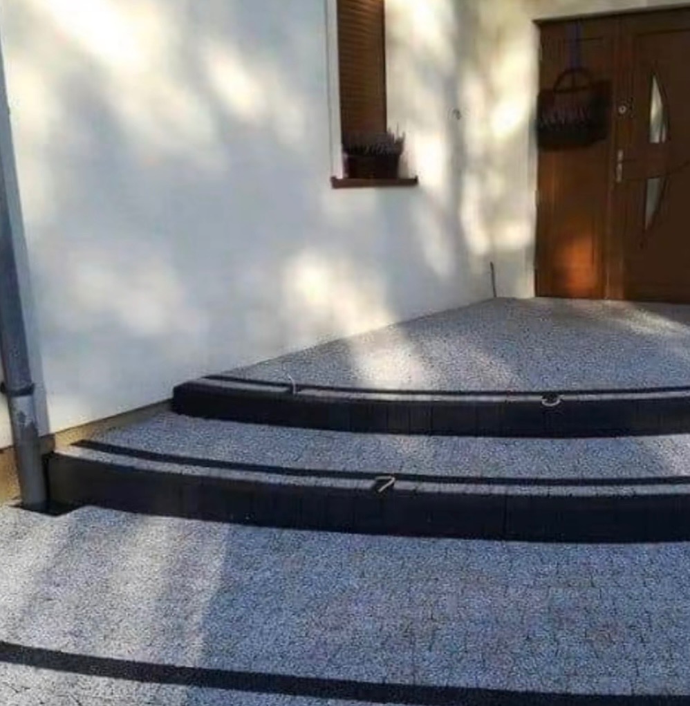 Schody wejściowe wyłożone kostką brukową w odcieniach szarości, z ciemnymi obrzeżami, prowadzące do drzwi domu. Widoczne lekkie cienie drzew na elewacji.