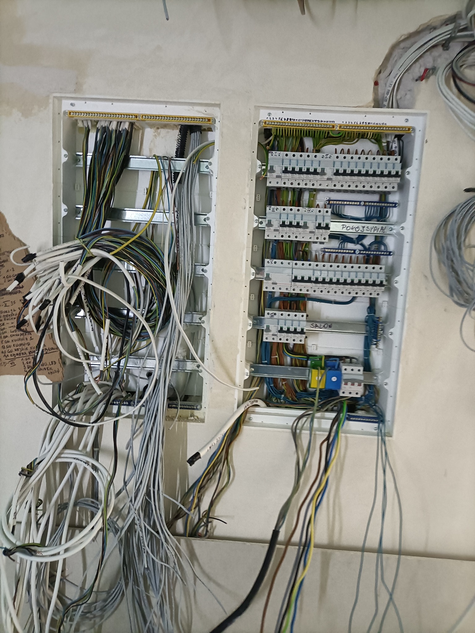 Dwie podtynkowe skrzynki elektryczne w trakcie montażu. Widoczne liczne przewody, listwy zaciskowe i zabezpieczenia. Na ścianie notatki. Instalacja w surowym stanie.