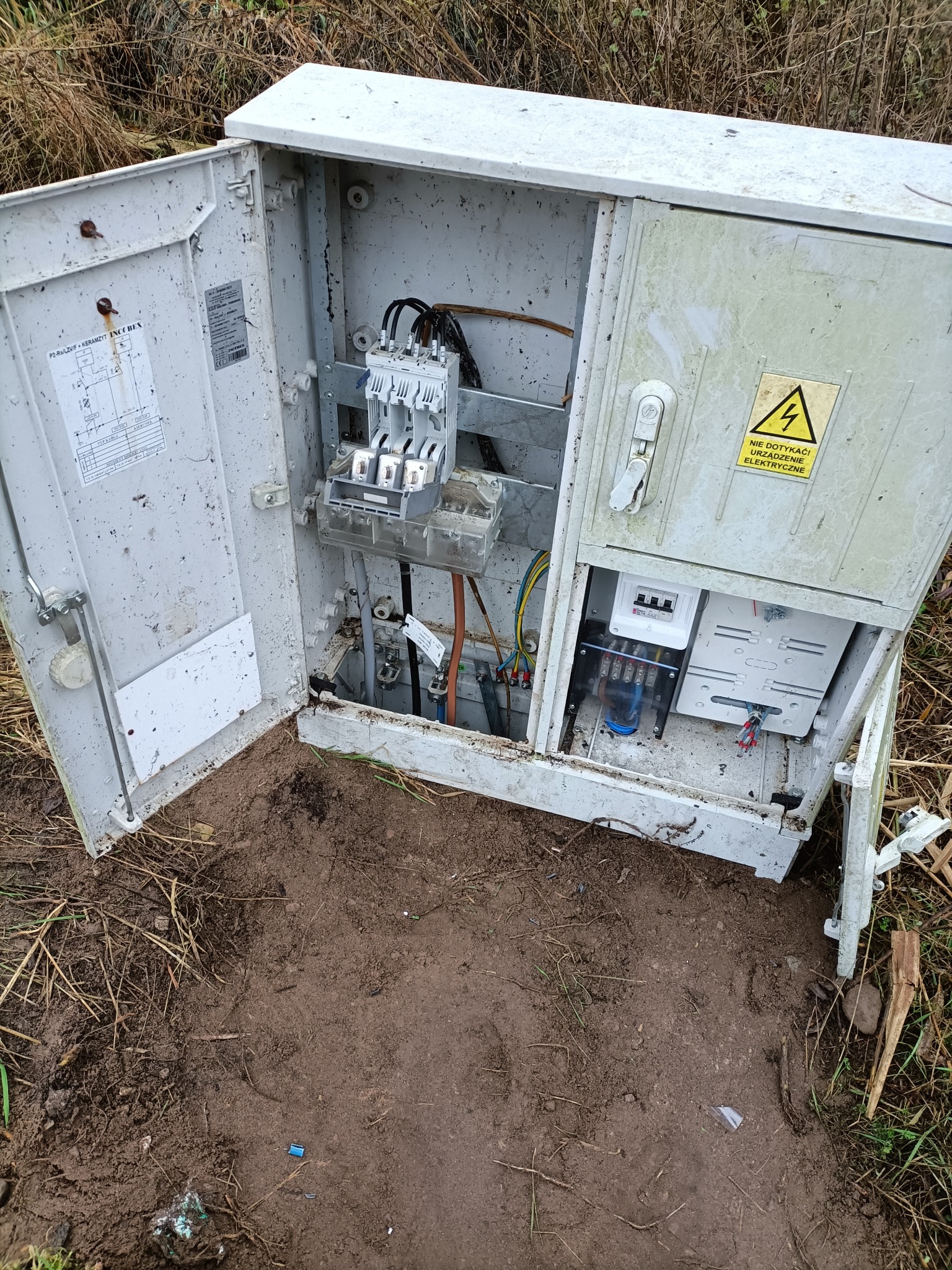 Otwarta, brudna skrzynka elektryczna na ziemi z widocznymi kablami, bezpiecznikami i licznikiem. Tabliczka ostrzegawcza z symbolem błyskawicy.