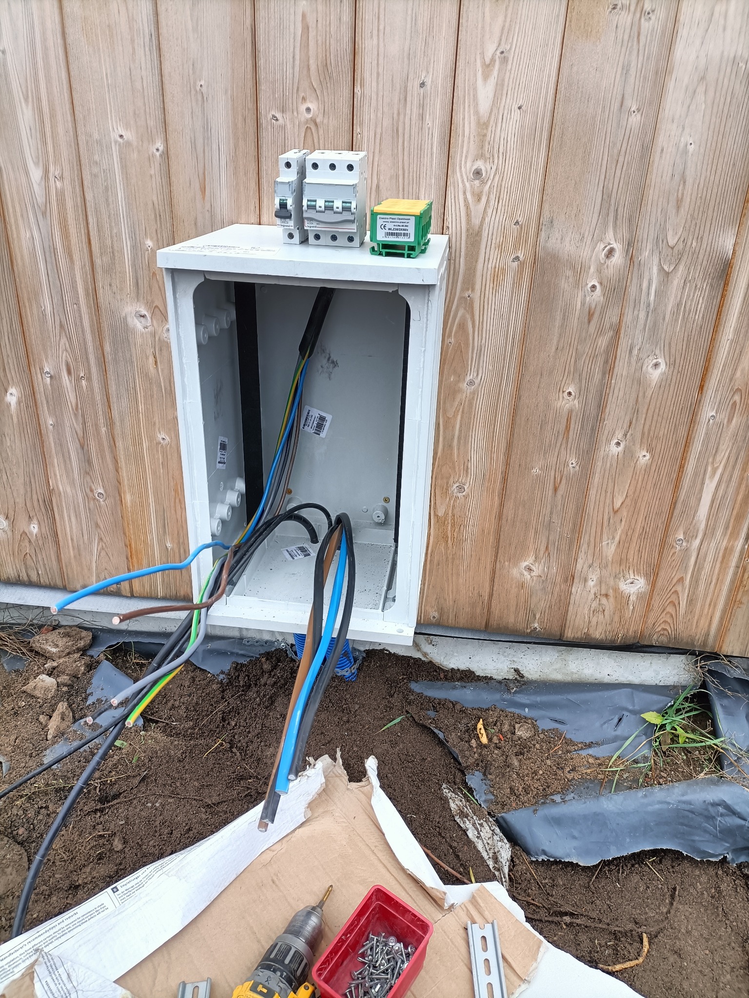 Otwarta skrzynka elektryczna z przewodami na tle drewnianej ściany, widoczne narzędzia i elementy montażowe. Realizacja przyłącza w trakcie prac ziemnych.