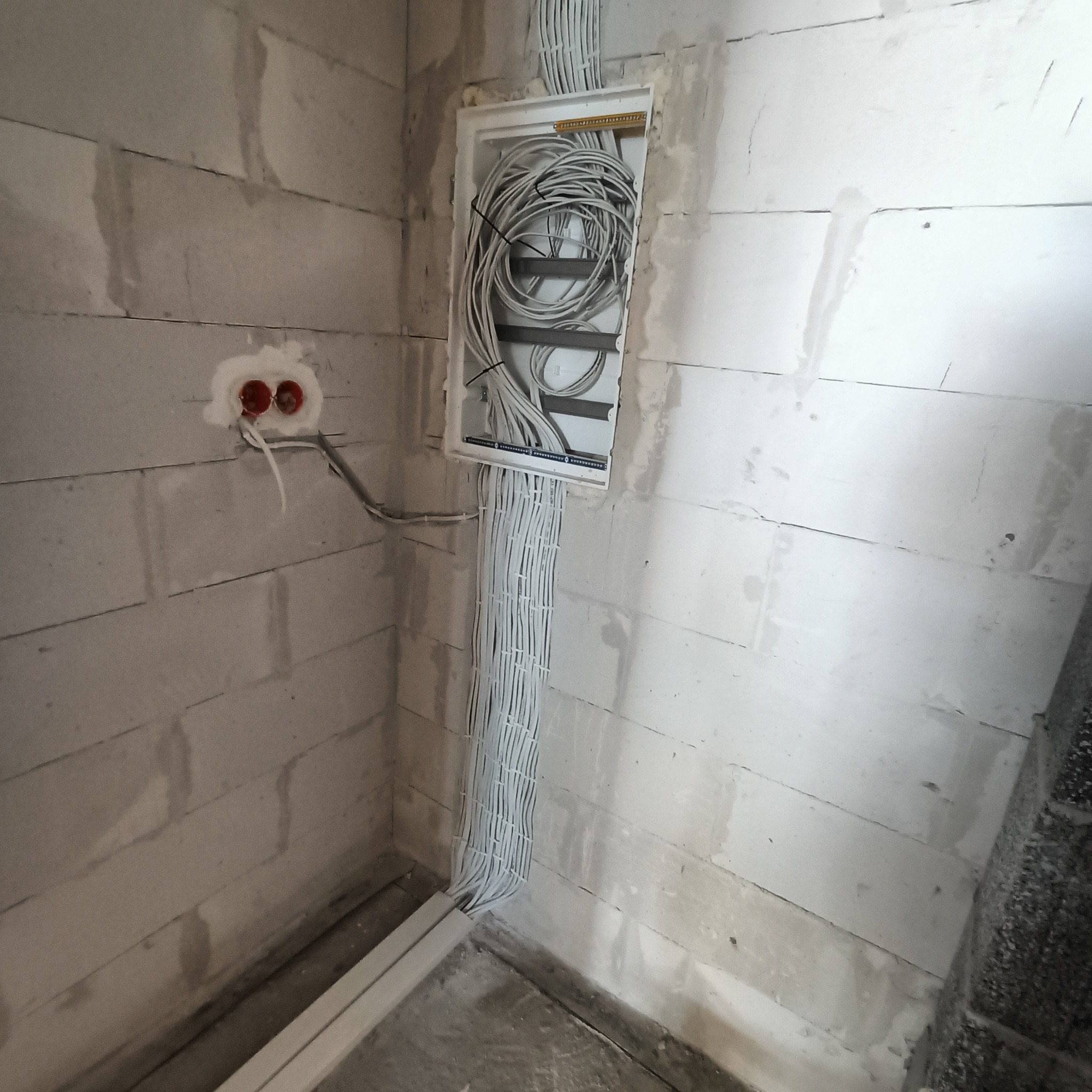 Wewnętrzna instalacja elektryczna w ścianie z białych bloczków, kable prowadzone w peszlach, puszka rozdzielcza z zapasem kabli. Widoczne gniazda elektryczne.