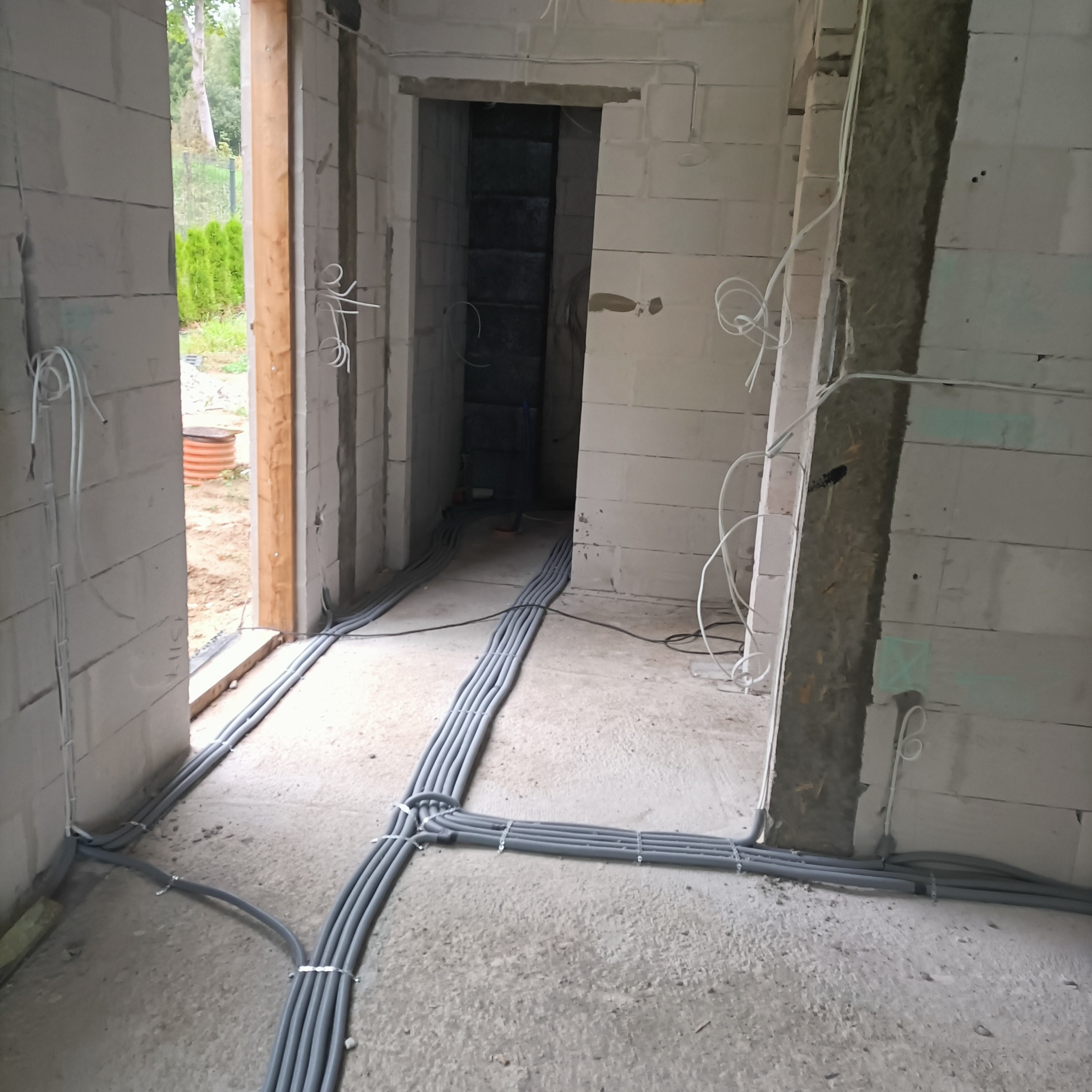 Szare rury instalacji elektrycznej ułożone równolegle na surowej posadzce wewnątrz budynku z bloków, widoczne wystające kable na ścianach.