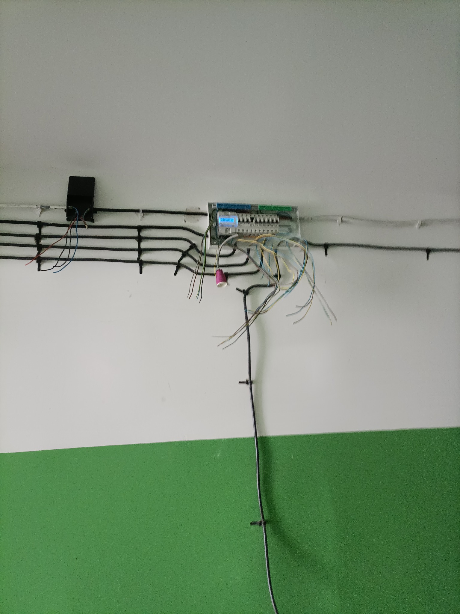 Fragment instalacji elektrycznej w budynku z przewodami, puszką i rozdzielnicą na biało-zielonej ścianie. Widoczne kable i elementy mocujące.