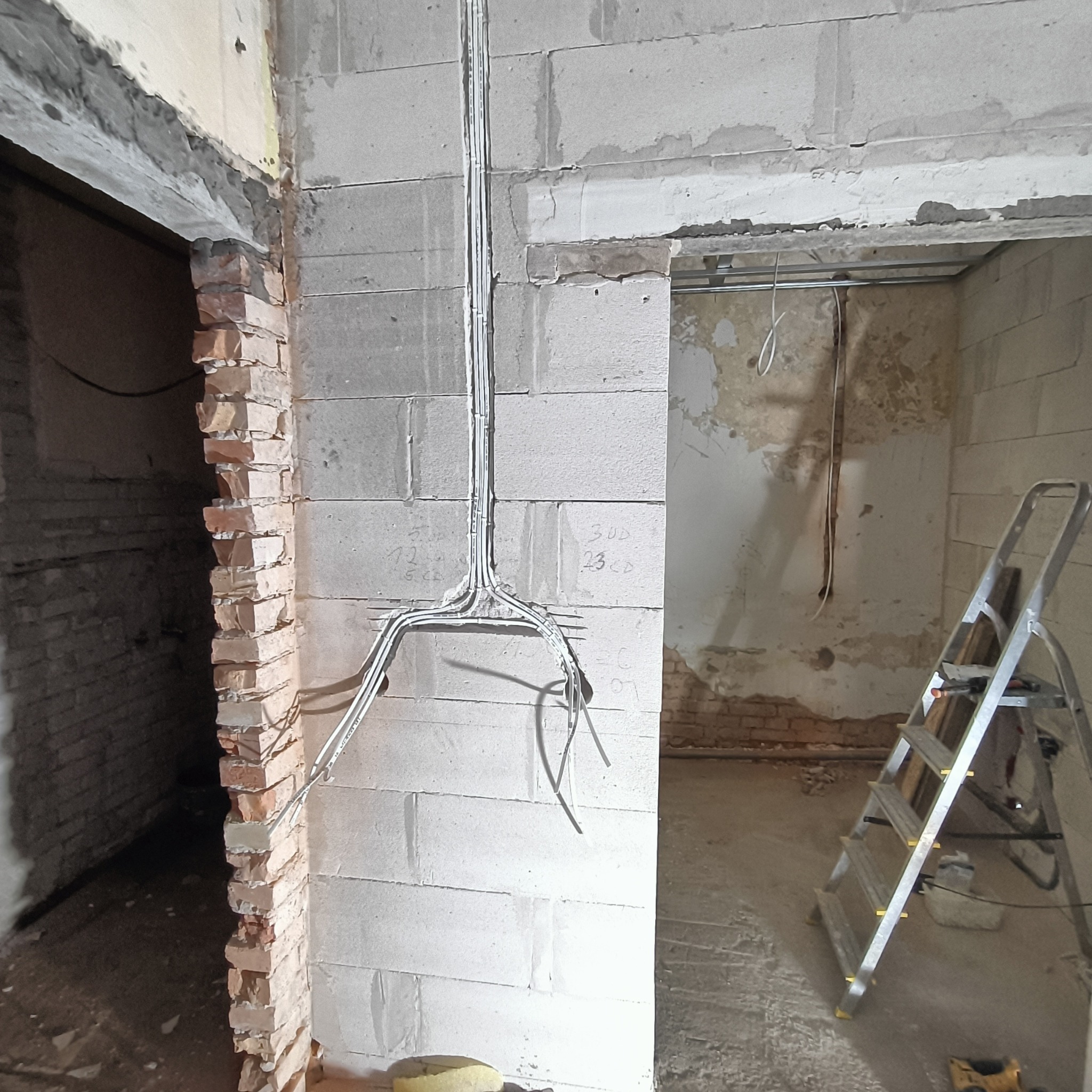 Ściana z instalacją elektryczną w trakcie remontu. Widoczne kable w bruzdach i drabina w przejściu. Surowy stan, brak tynków. Odsłonięta cegła w drugim przejściu.