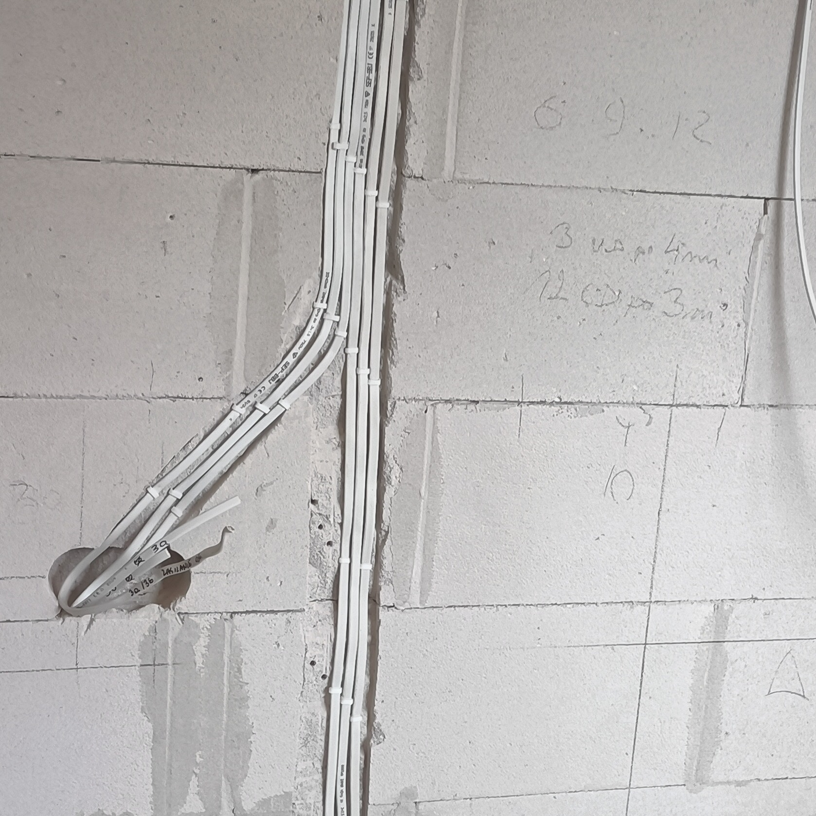 Białe kable elektryczne ułożone w bruzdach ściany z betonu komórkowego, z widocznymi oznaczeniami i notatkami na ścianie. Etap prac instalacyjnych.