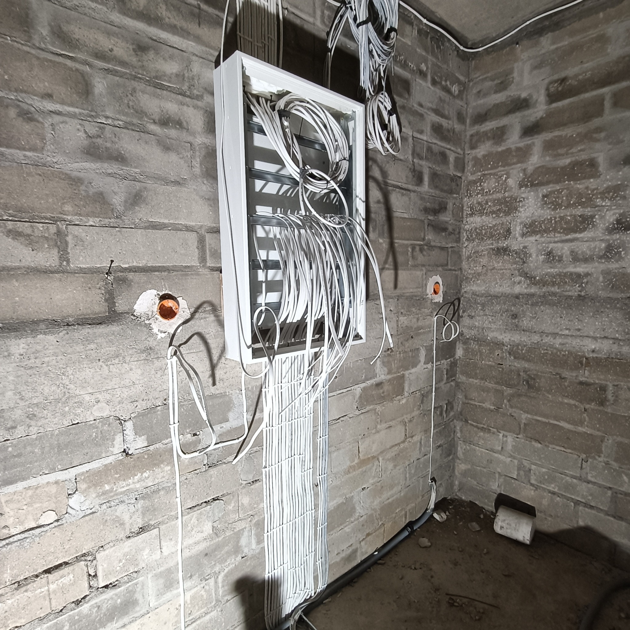 Rozdzielnia elektryczna z licznymi przewodami na tle surowej, ceglanej ściany. Widoczne otwory w ścianie z pomarańczowymi wkładami. Przewody ułożone pionowo.
