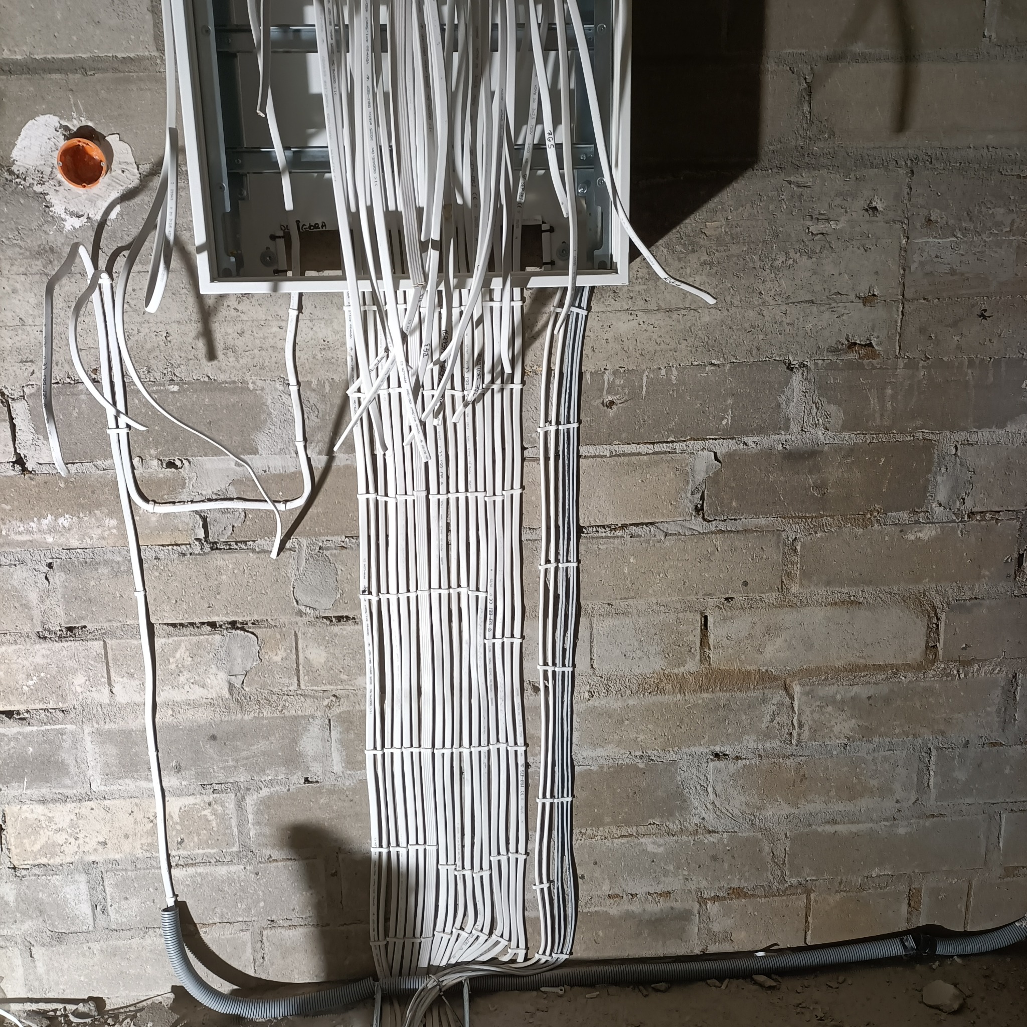 Rozdzielnica elektryczna z licznymi przewodami na surowej, ceglanej ścianie. Kable uporządkowane w wiązki, część w peszlu. Widoczny fragment instalacji w trakcie montażu.