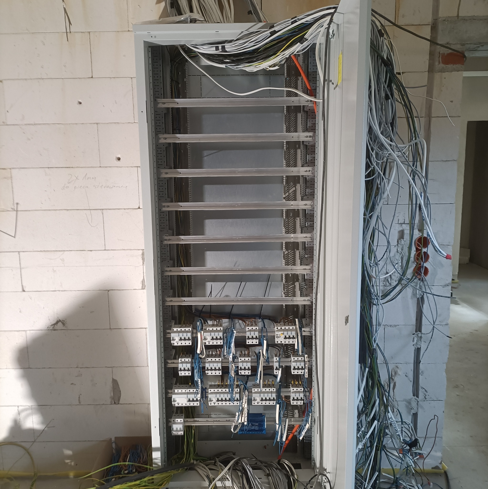 Rozdzielnica elektryczna w trakcie montażu w surowym wnętrzu. Widoczne liczne przewody, zabezpieczenia i elementy montażowe. Ściany z białego betonu komórkowego.