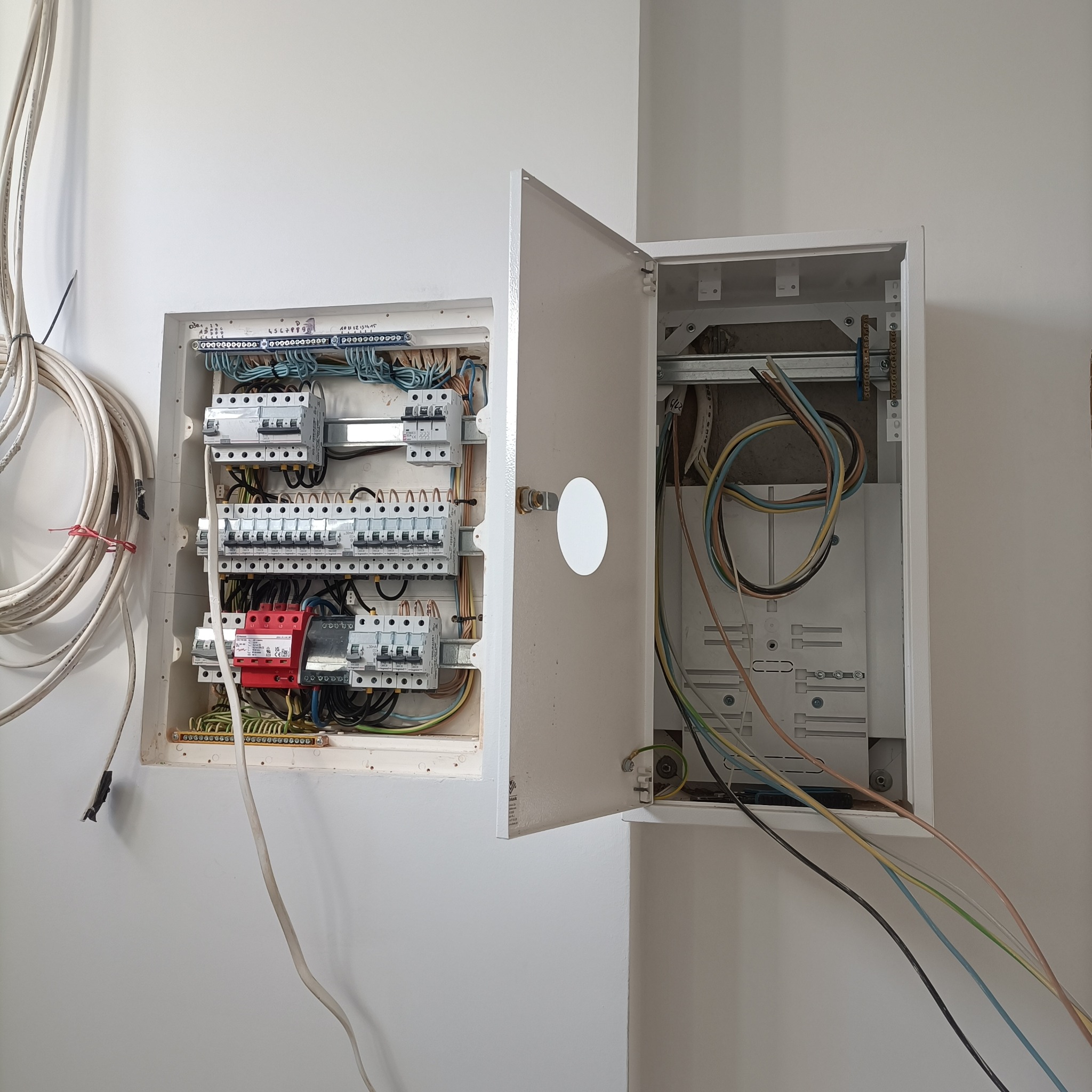 Otwarta skrzynka elektryczna z zainstalowanymi bezpiecznikami i przewodami na białej ścianie. Widoczne kable zwisające z boku. Instalacja w trakcie realizacji.