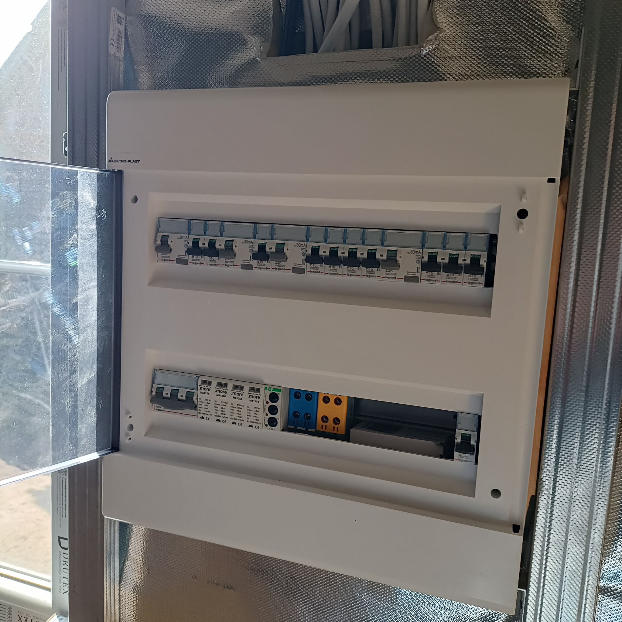 Zamknięta skrzynka elektryczna Elektro-Plast z zainstalowanymi bezpiecznikami Legrand i .more, zamontowana na ścianie z izolacją. Widoczne przewody.