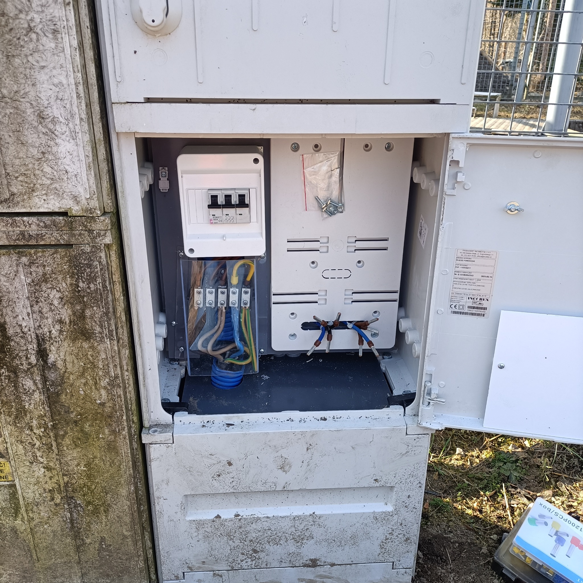 Otwarta skrzynka elektryczna z widocznymi przewodami, bezpiecznikami i elementami złącznymi. Widoczne ślady użytkowania na obudowie.