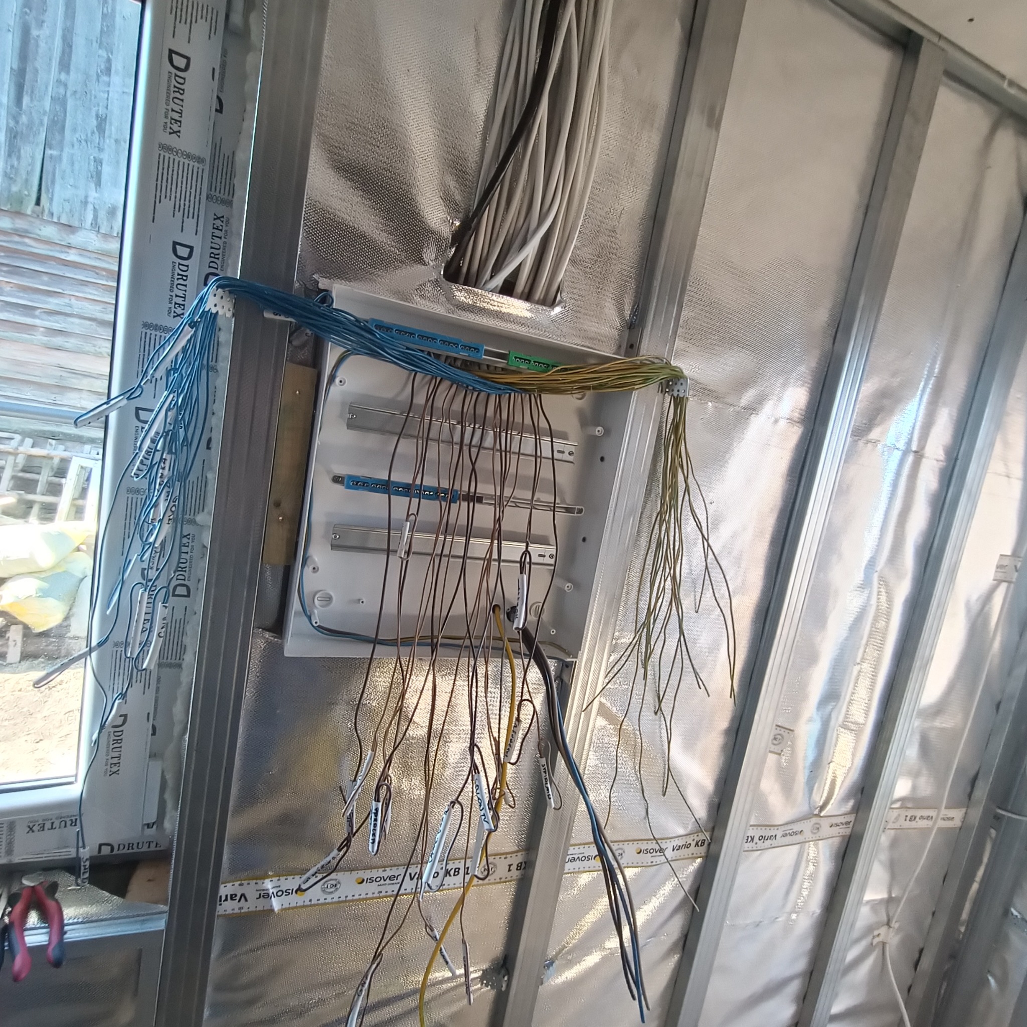 Rozdzielnica elektryczna z podłączonymi przewodami w budynku w trakcie budowy, widoczne kable oznaczone etykietami, ściany wyłożone folią izolacyjną.
