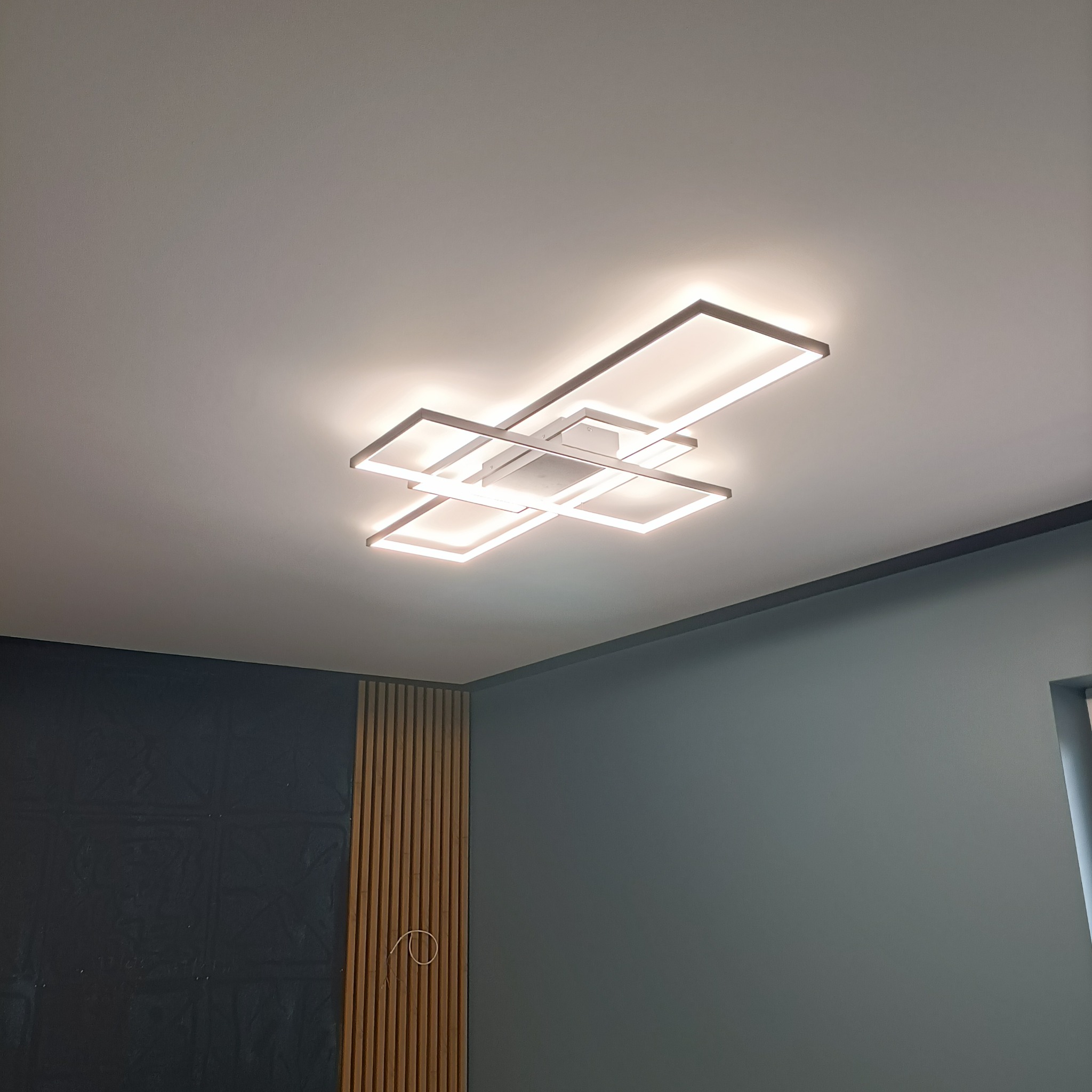 Nowoczesna lampa sufitowa LED w kształcie połączonych kwadratów, oświetlająca wnętrze z ciemnymi ścianami i drewnianymi panelami. Minimalistyczny design.